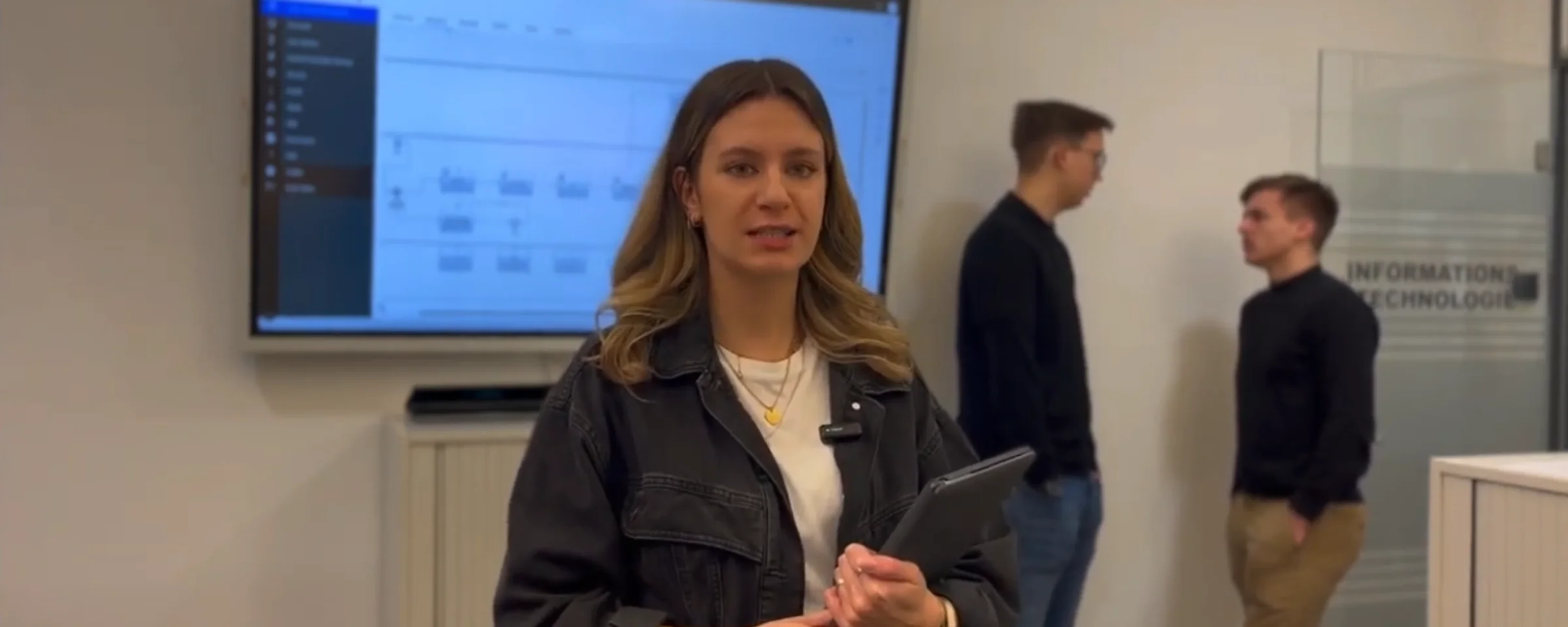 Eine Frau steht mit einem Tablet in einem Büro, im Hintergrund ein Bildschirm mit Grafiken. Nolte Küchen.