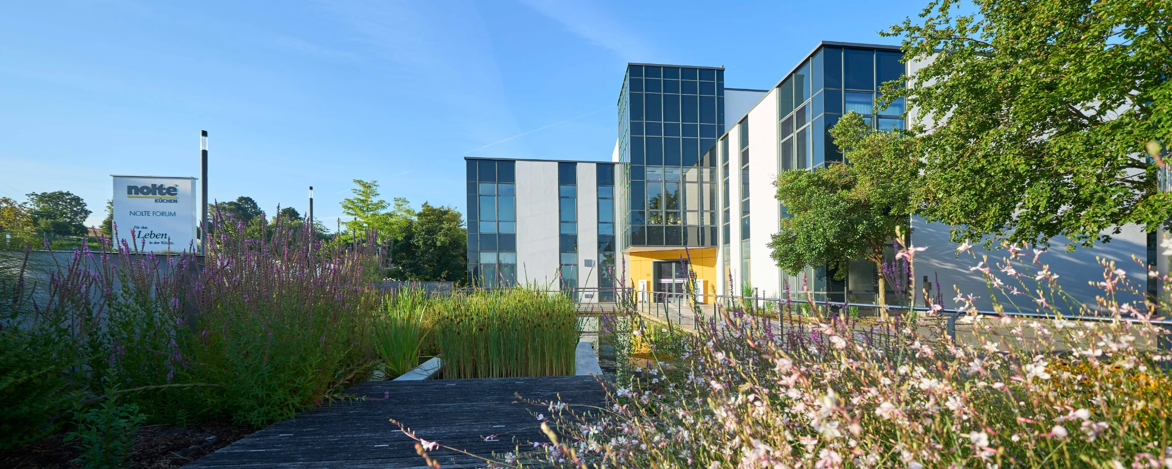 Ein modernes Gebäude umgeben von Blumen und Bäumen bei blauem Himmel, Nolte Küchen.