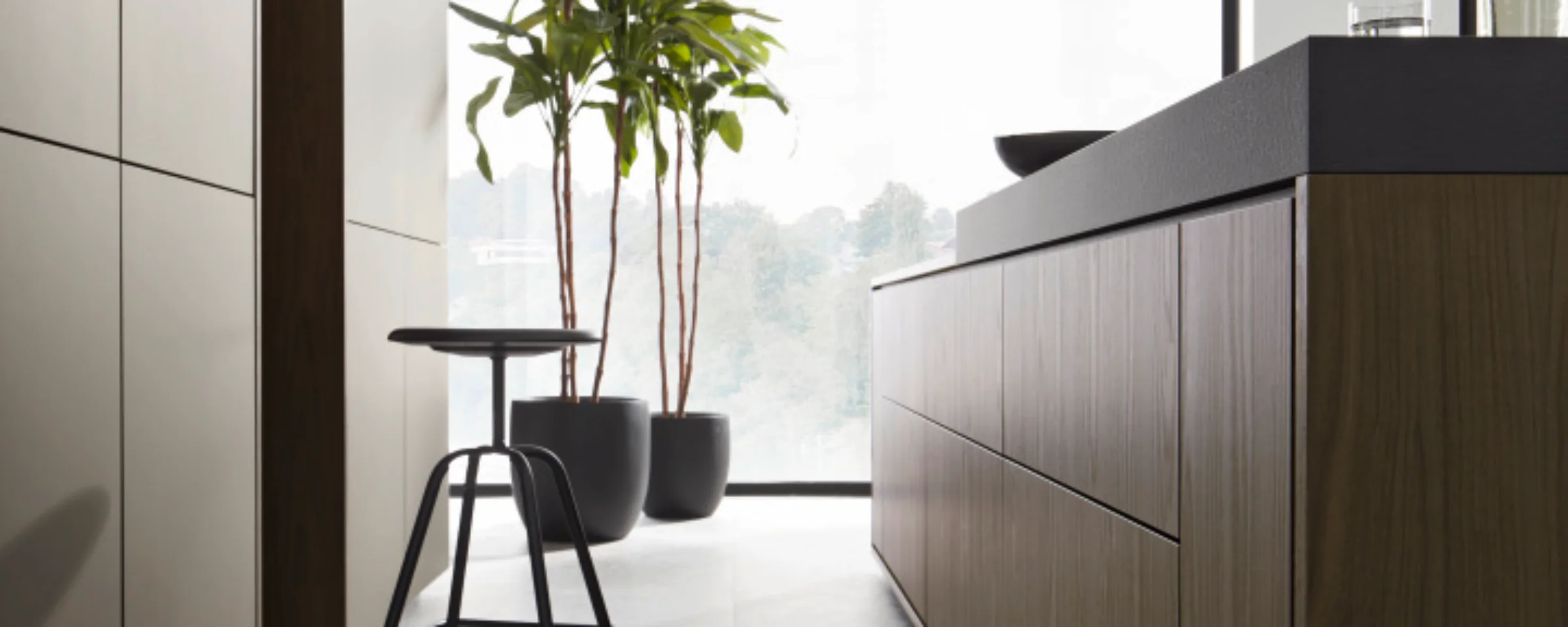 Moderne Küche mit Kochinsel und Barhocker, im Hintergrund Pflanzen vor einem großen Fenster. Nolte Küchen.