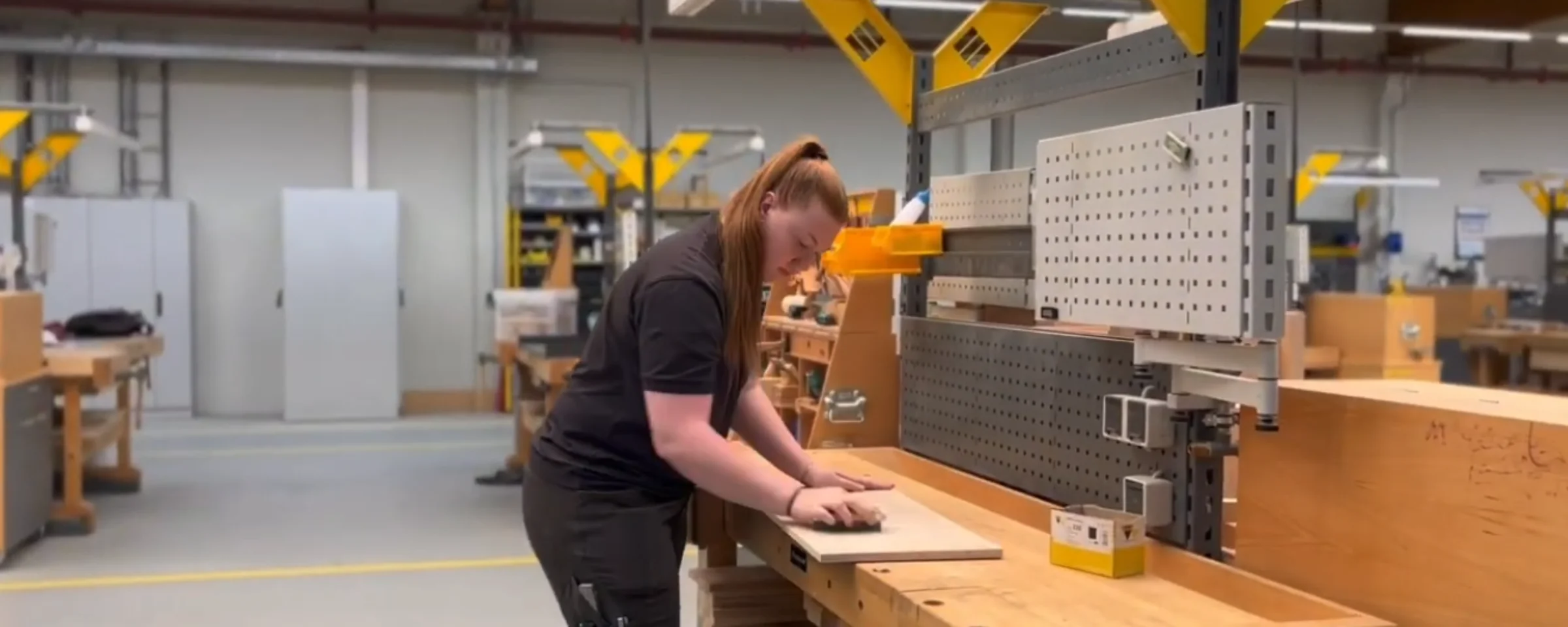 In einer Werkstatt arbeitet eine Person an einem Holztisch mit Werkzeugen; im Hintergrund sind weitere Arbeitsplätze zu sehen.