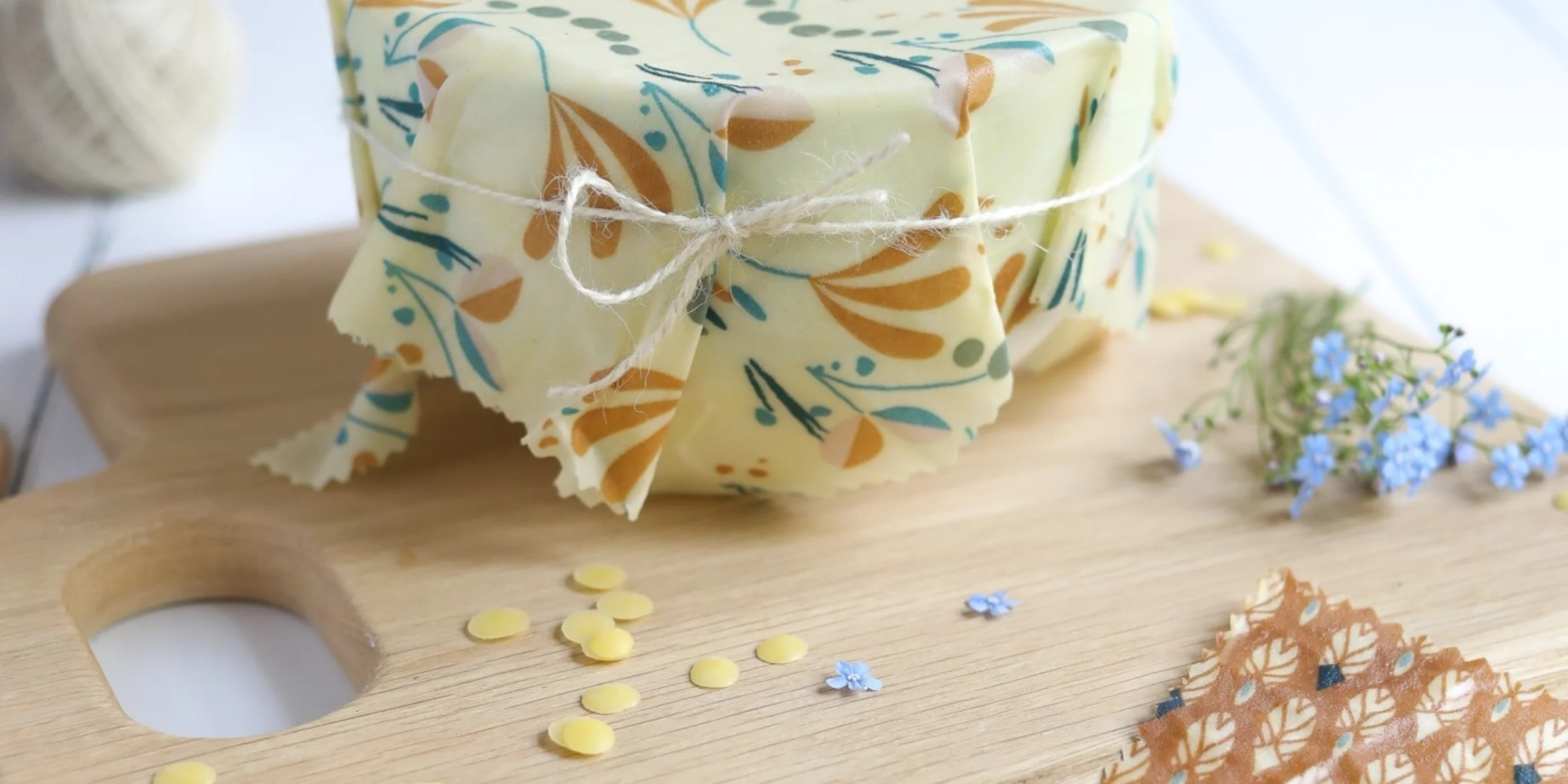 Eine Schale mit Bienenwachstuch auf einem Holzbrett, umgeben von Blumen und gelben Wachsstücken.