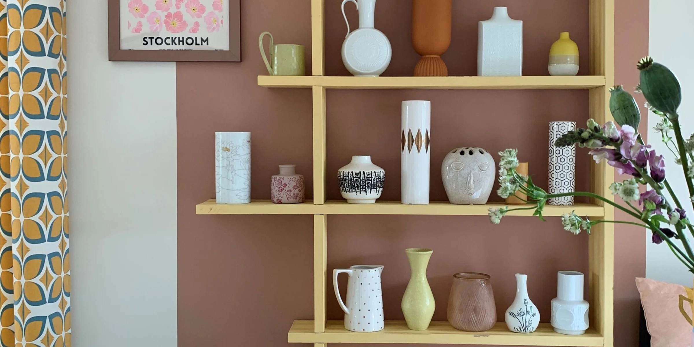 Ein Regal mit Vasen und Dekor vor einer rosa Wand, mit einem Kissen und Magazin im Vordergrund. Nolte Living.