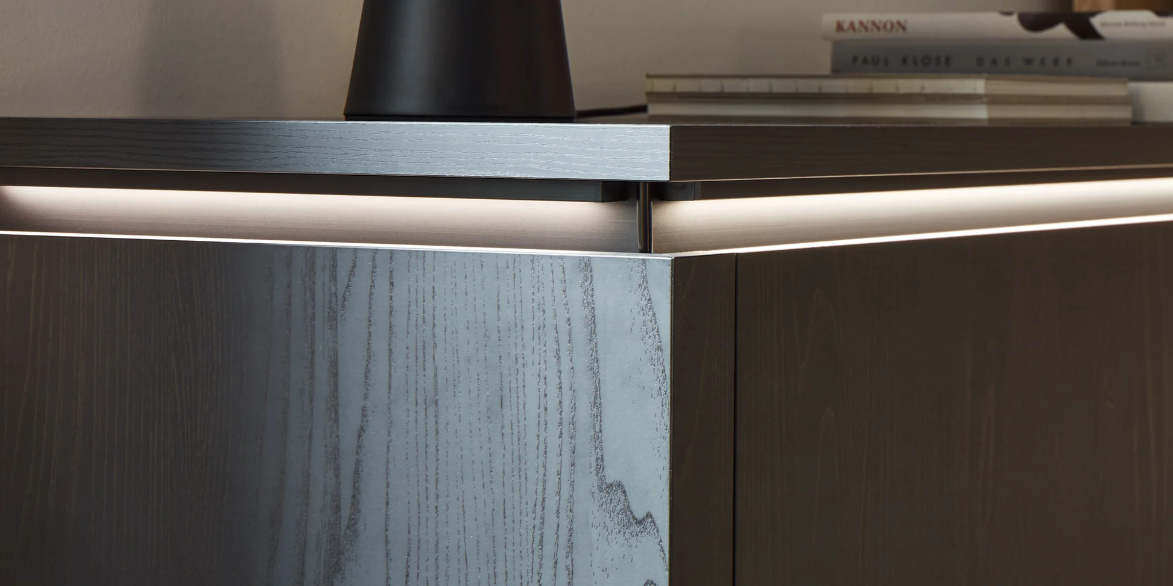 Ein modernes Sideboard mit dunklem Holzdesign und Beleuchtung, Bücher und Dekoration darauf, von Nolte Living.