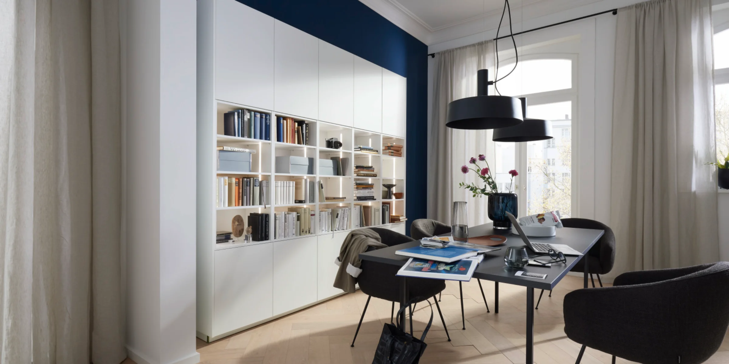 Ein modernes Wohnzimmer mit einem eleganten Bücherregal, Tisch und Stühlen, passend zu Nolte Living.