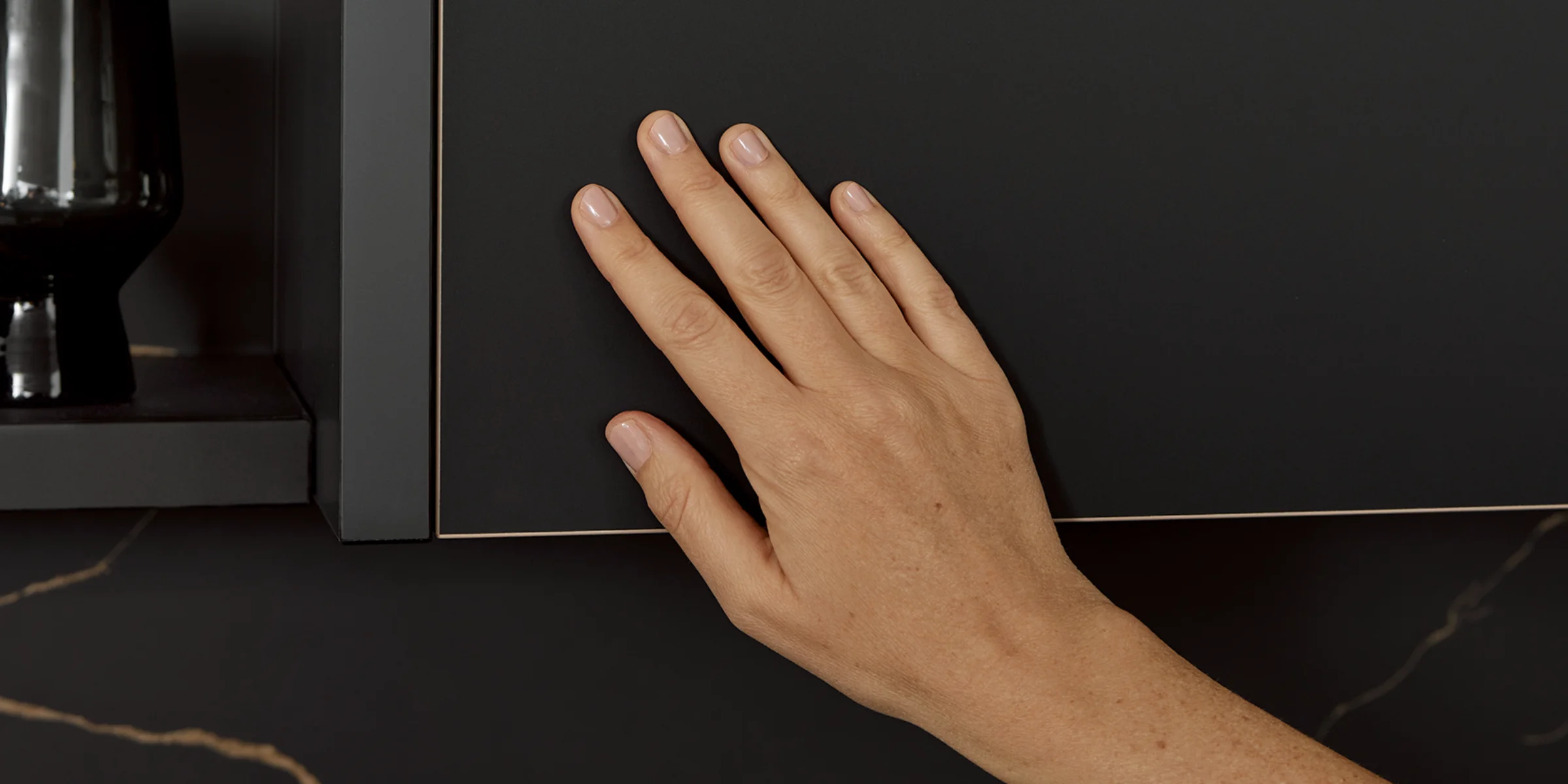 Hand berührt eine matte schwarze Küchenfront FLAIR mit feiner Kante in Messingoptik, die eine Anti-Fingerprint-Oberfläche besitzt.