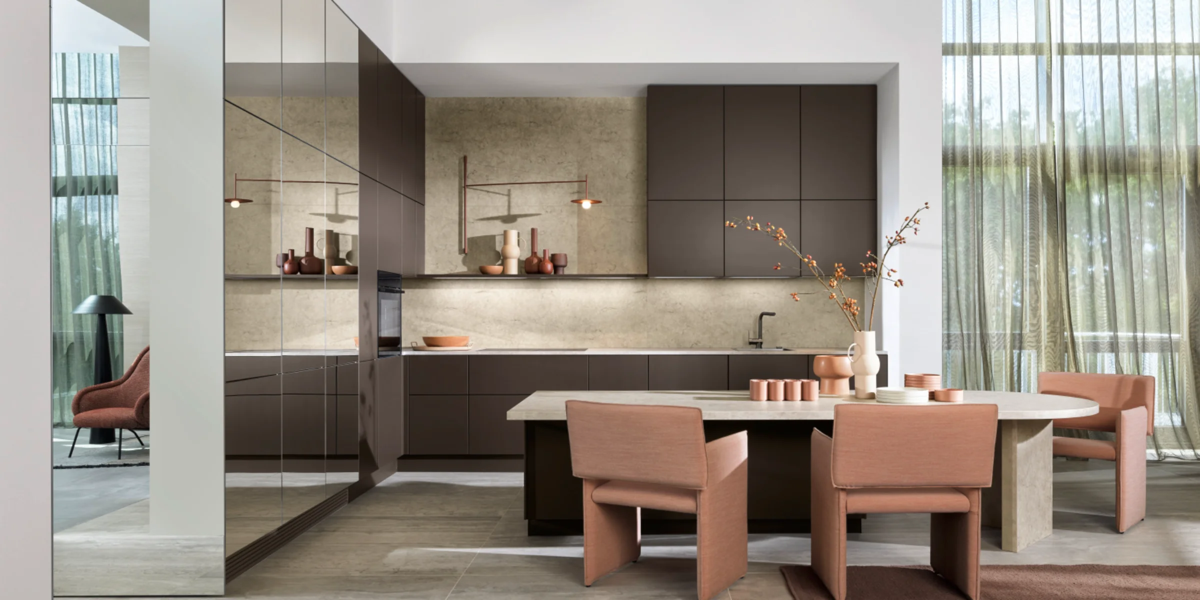 Cuisine Nolteneo moderne avec façade neoMIRROR en aspect miroir et façade Soft Lack en brun sépia. Armoires sans poignées, plan de travail couleur pierre et table à manger ronde avec chaises rembourrées dans une ambiance élégante et baignée de lumière.