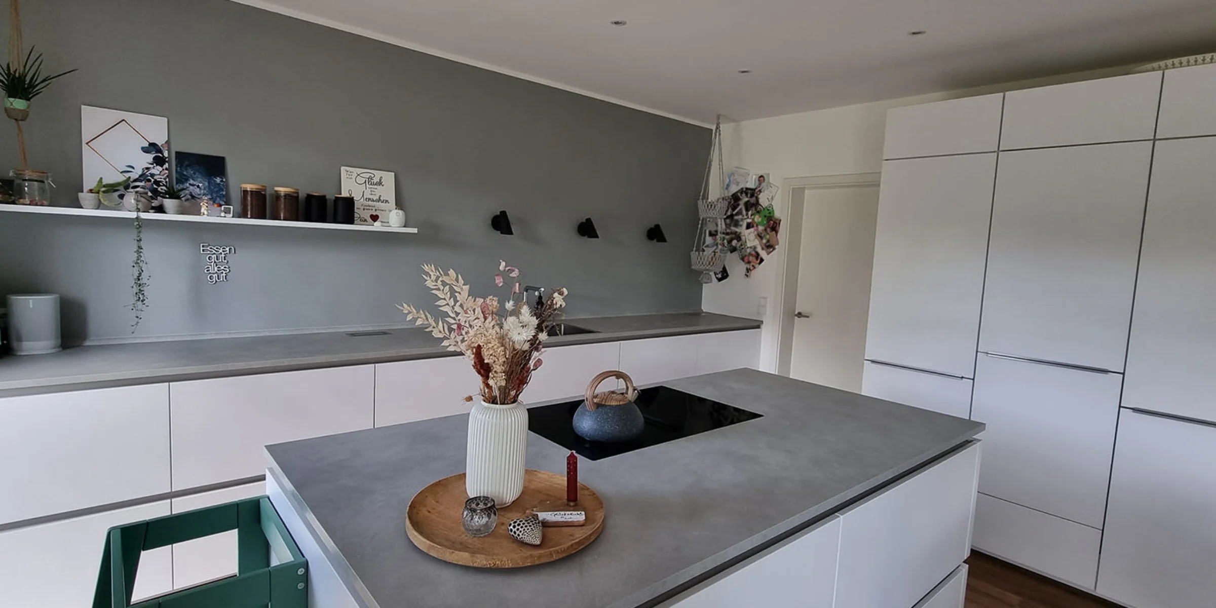 Modern, minimalist Nolte kitchen of a Nolte Küchen employee with a concrete worktop and fronts from the PLUS range in Arcticweiß. In the centre there is a kitchen island with an induction hob and a vase with dried flowers. On the wall hangs an open shelf with decoration, glasses and a mint-green coffee machine. A magnetic wall with photos and hanging baskets gives the kitchen a personal touch. The kitchen appears tidy, stylish and functional.