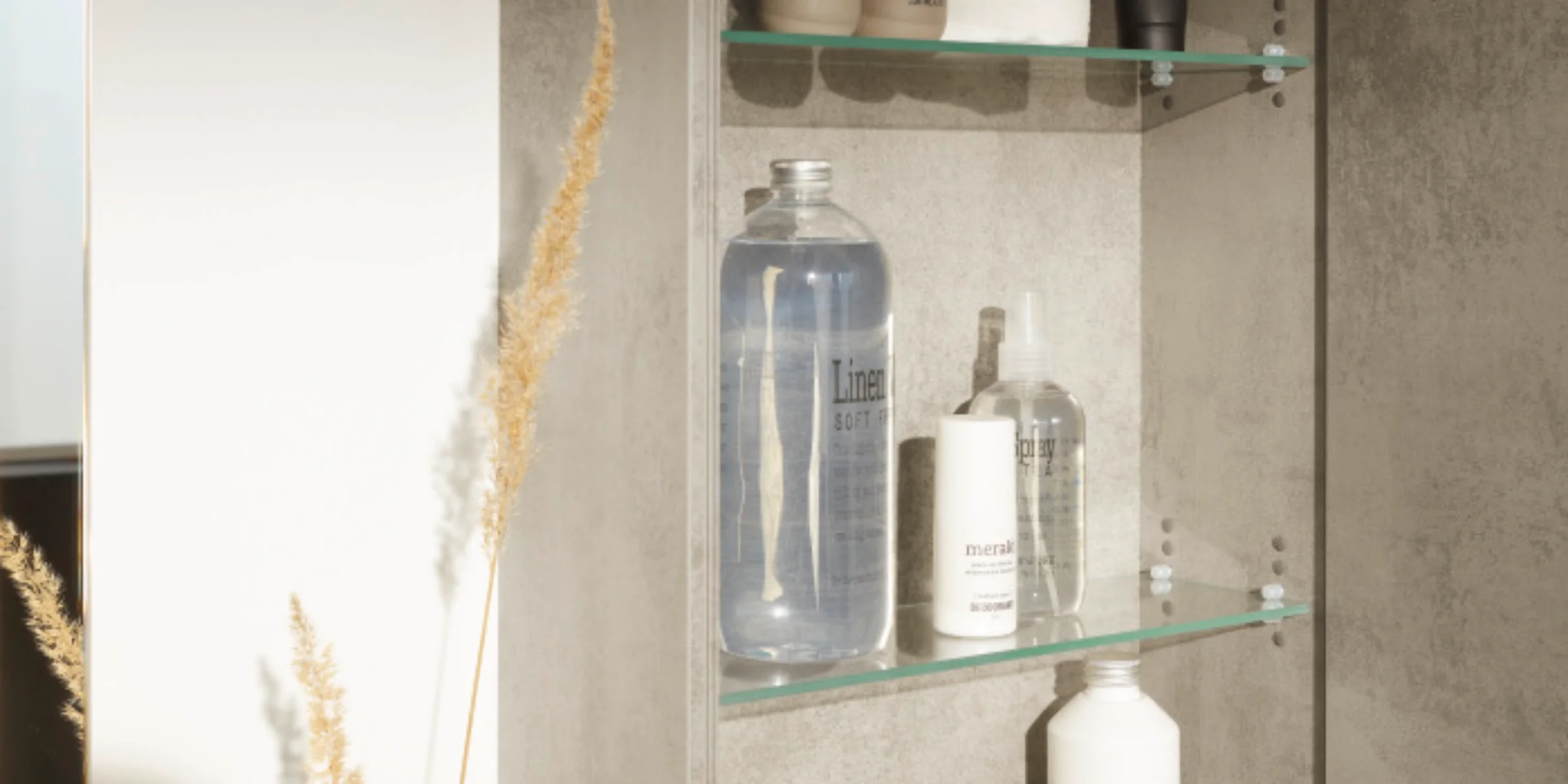 Ein geöffneter Badezimmerschrank mit Pflegeprodukten, neben einer Vase mit Gräsern. Nolte SPA.