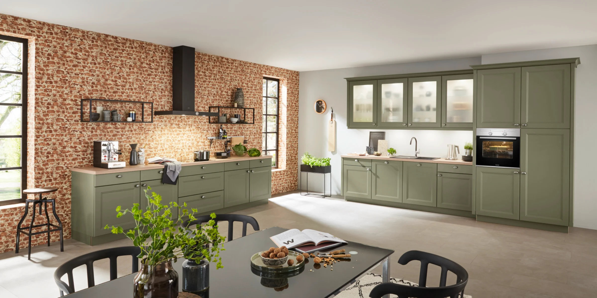 Landhausküche von Nolte mit der Front WINDSOR LACK in Olive softmatt. Die natürliche Farbgebung wird durch eine Arbeitsplatte in Steineiche ergänzt. Schwarze Wandregale im Industrial Style setzen moderne Kontraste zur klassischen Rahmenoptik der Fronten. Die Küche vereint ländliche Gemütlichkeit mit urbaner Klarheit – funktional, wohnlich und stilvoll.