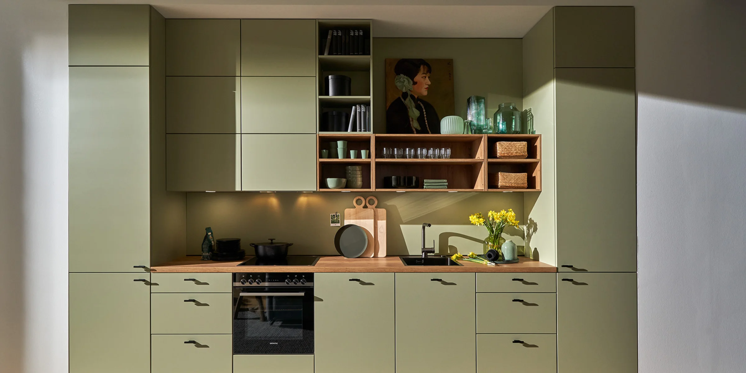 Stilvolle Nolte Küche mit der Front SOFT LACK in Avocado softmatt. Die grifflose Küchenzeile kombiniert sanftes Grün mit einer warmen Holzarbeitsplatte und offenen Regalen. Die wohnliche Atmosphäre wird durch dekorative Elemente, Pflanzen und natürliche Materialien unterstrichen – eine moderne Küche mit frischer Farbgebung und klarer Linienführung.