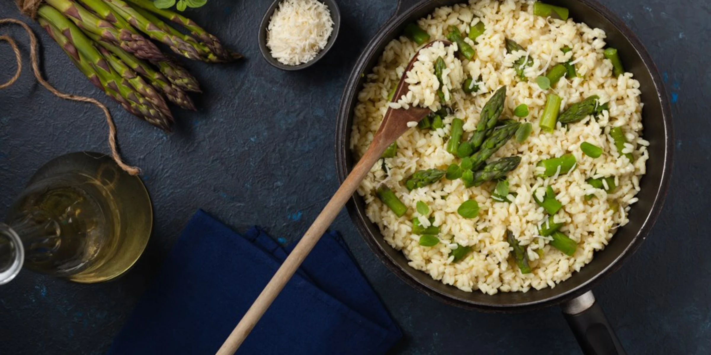 Eine Pfanne mit Spargelrisotto neben frischen Spargelstangen und einem Glas Weißwein auf dunklem Untergrund.