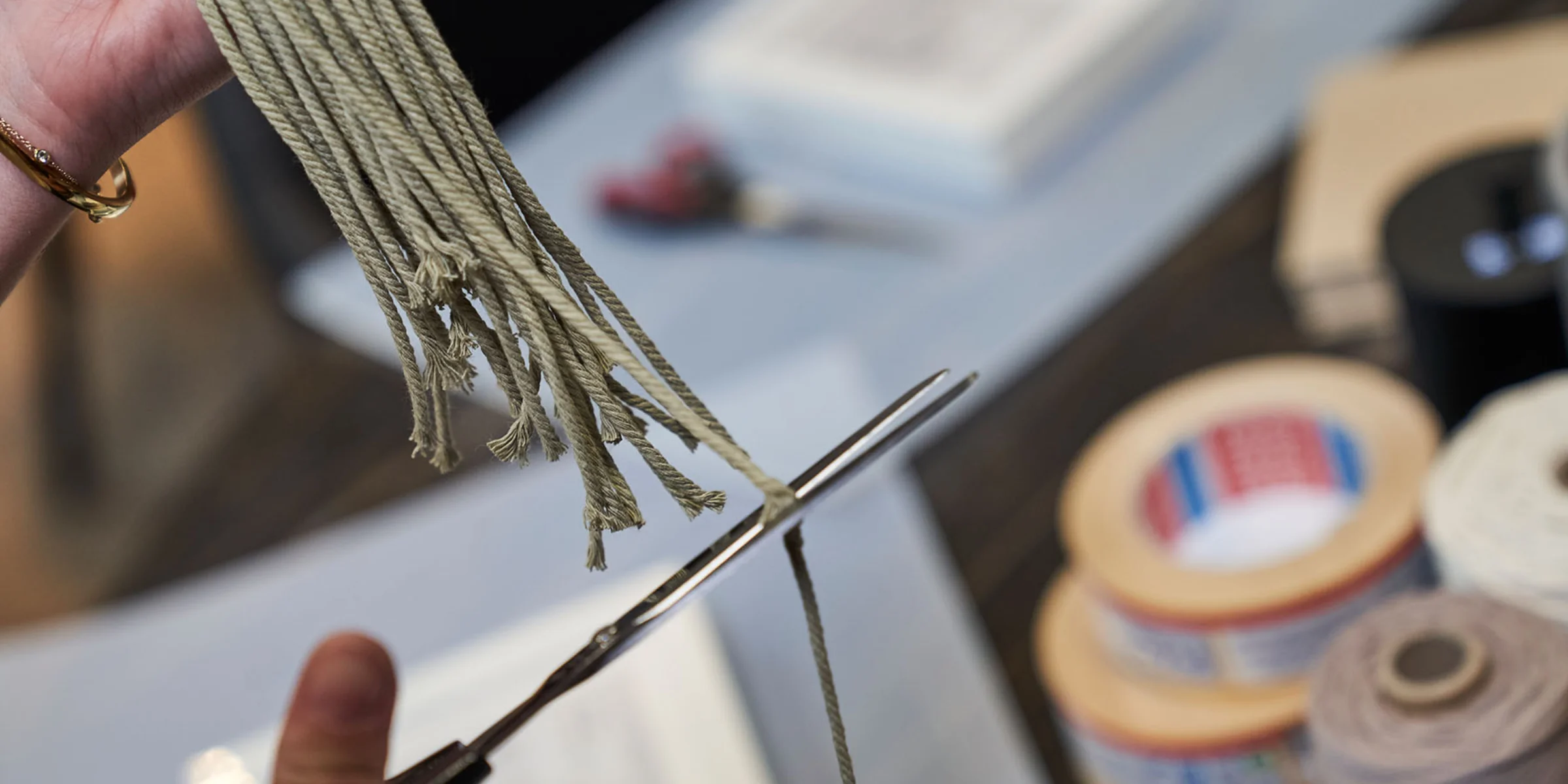 Eine Person schneidet grüne Fäden mit einer Schere, im Hintergrund sind Klebebänder und Papier zu sehen.