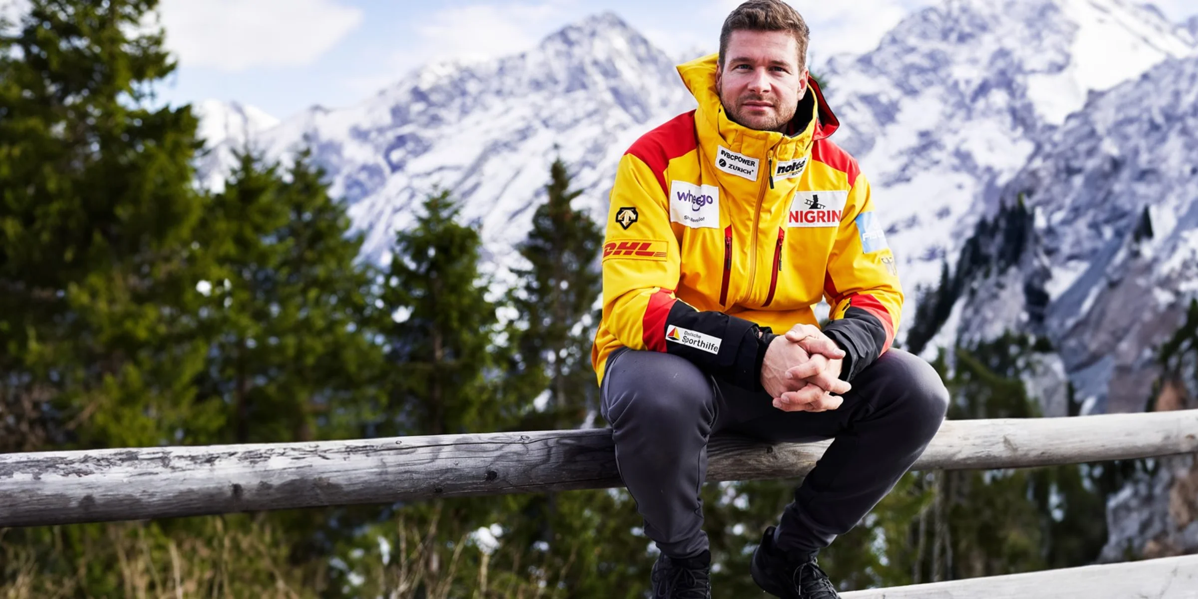 Ein Mann in einer gelben Jacke sitzt vor schneebedeckten Bergen auf einem Holzzaun.