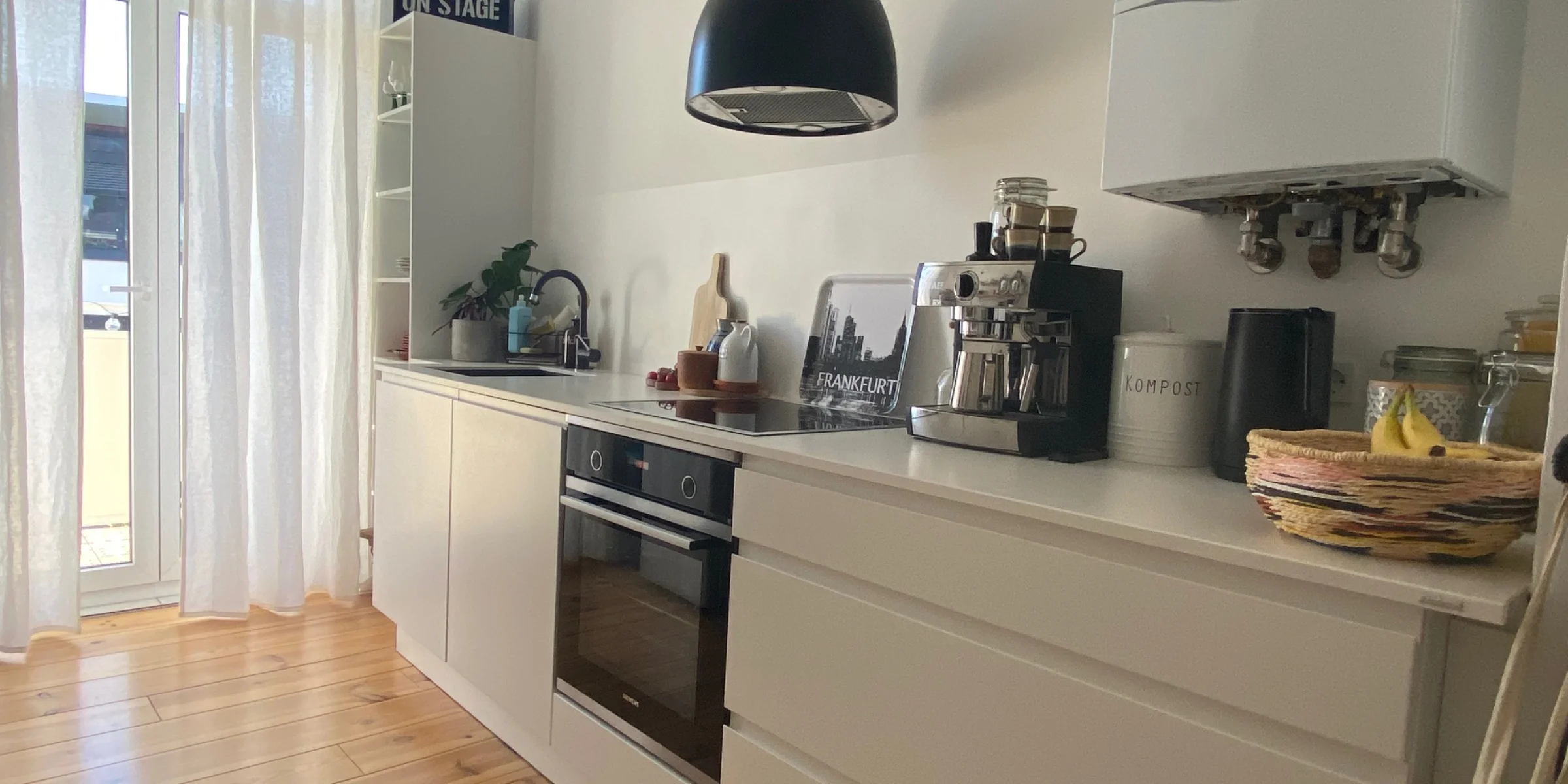 The modern Nolte kitchen featuring the SIGMA LACK range is located in a bright room with large windows and floor-length white curtains. The handleless white cabinets and drawers harmonize with the warm wooden floor. A black built-in oven with cooktop is centrally positioned, next to it a sink with a dark faucet. On the worktop, installed by the customer, stand an espresso machine, a black water jug and a compost container. A black pendant lamp adds a modern accent. Personal details such as an "ON STAGE" sign and a Frankfurt skyline tray complete the ambience.