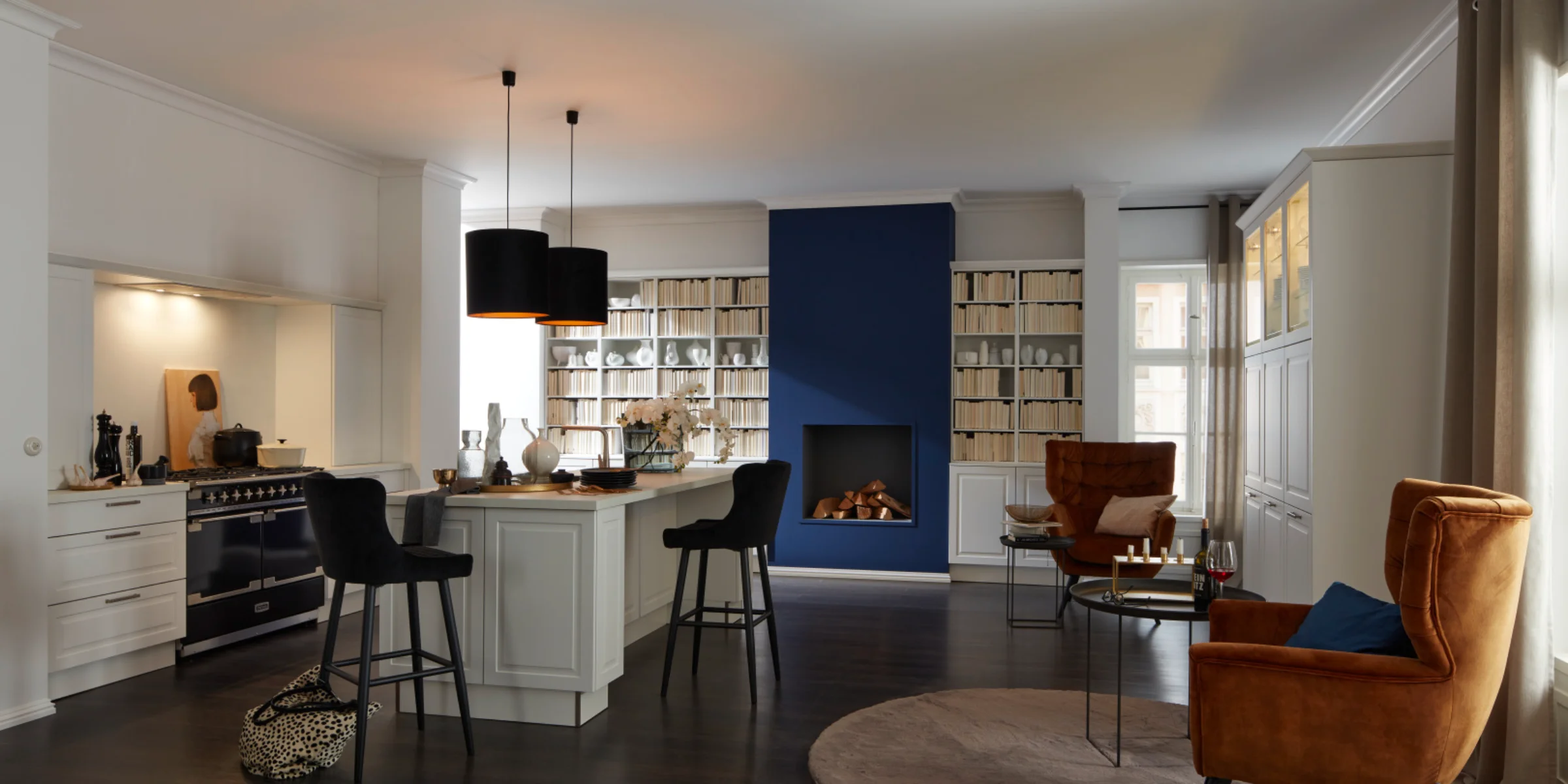 Gemütliche Nolte Küche im Landhausstil mit der Front PORTO Weiß softmatt. Die Küche überzeugt mit klassischer Rahmenoptik, einer schwarzen Landhauskochstelle und einem Kochinselbereich mit Barhockern. Helle Fronten harmonieren mit dem dunklen Holzboden, während ein integrierter Wohnbereich mit Kamin, Bücherregalen und samtigen Lounge-Sesseln in Cognac für ein elegantes, wohnliches Ambiente sorgt.