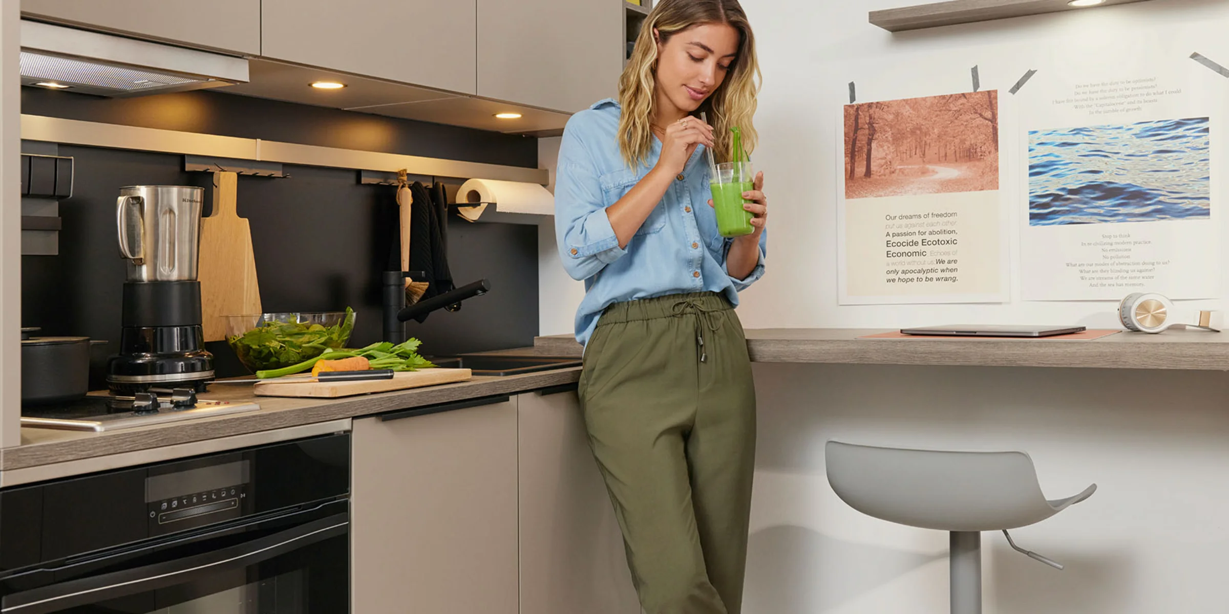 Junge Frau in moderner, kompakter Küchenzeile mit Frontenprogramm PLUS in Lava in einem Micro Appartment mit Smoothie in der Hand, vor einer Arbeitsfläche mit frischem Gemüse und Mixer.