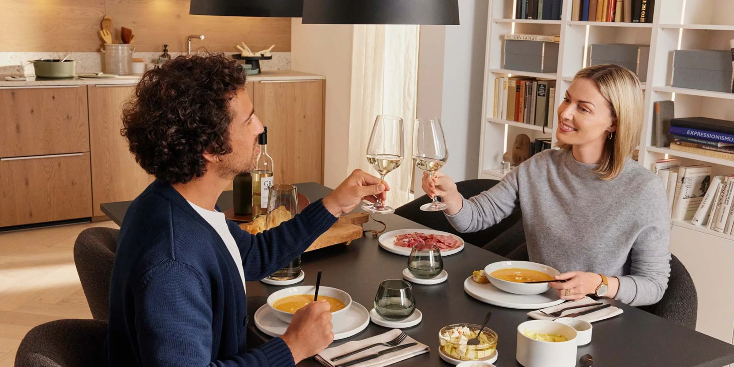 Ein Paar isst in einer modernen Küche und stößt mit Weingläsern an. Nolte Küchen.