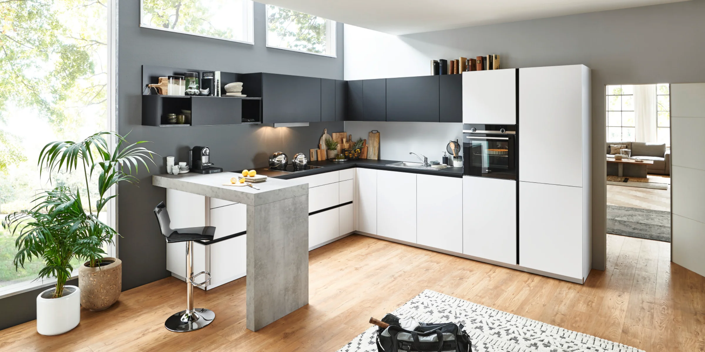 Moderne Nolte Küche in L-Form und Tresen mit FEEL Fronten in Arcticweiß softmatt und PLUS Fronten in Schwarz softmatt. Die grifflose Gestaltung wird durch eine markante Theke in Beton-Optik ergänzt. Die helle, offene Raumwirkung wird durch viel Tageslicht und natürliche Materialien wie Holzfußboden und Pflanzen unterstützt – ein stilvoller, funktionaler Raum zum Kochen und Leben.