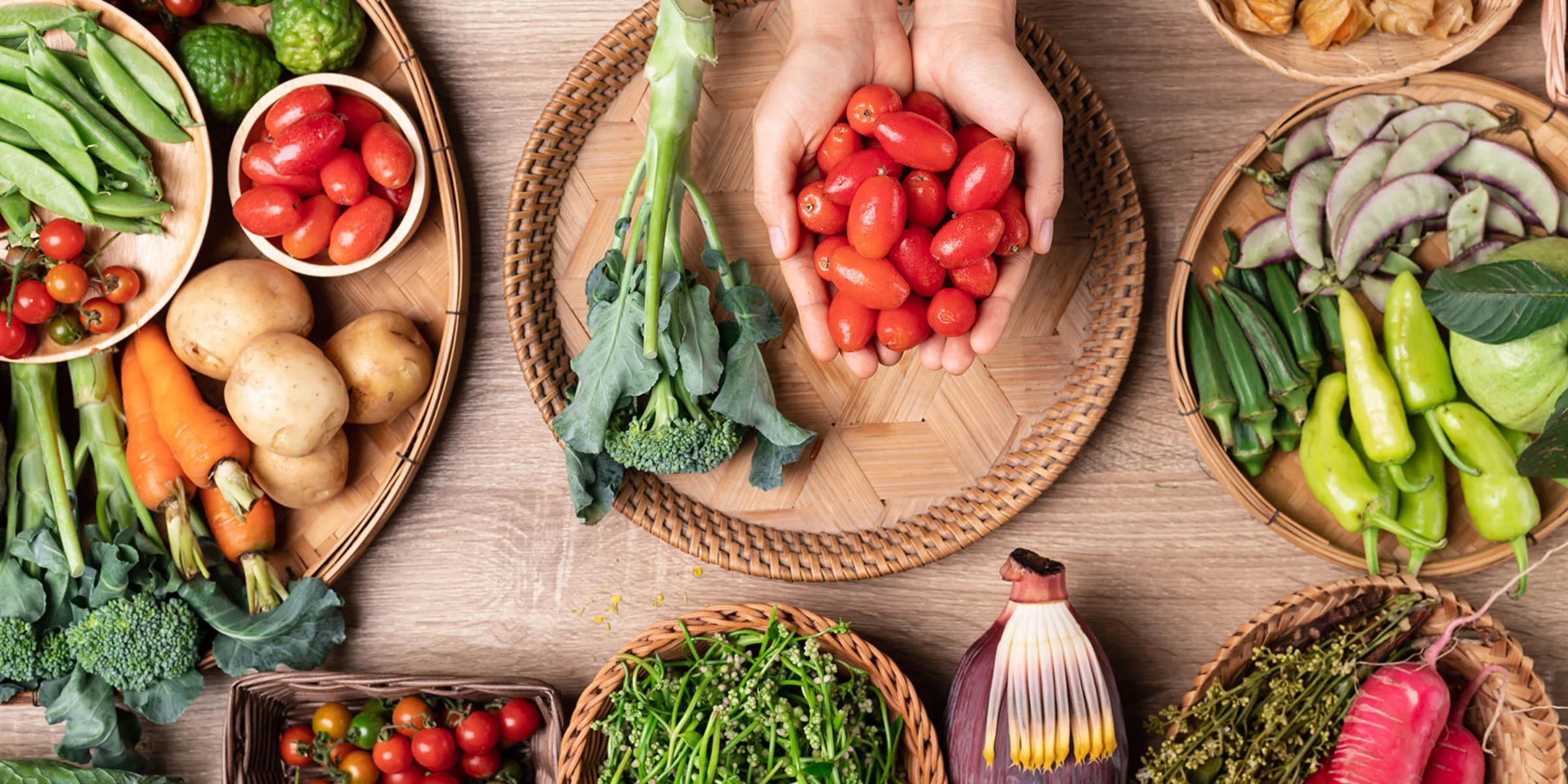 Verschiedene frische Gemüsesorten liegen auf einem Tisch, in der Mitte hält jemand Kirschtomaten in den Händen.