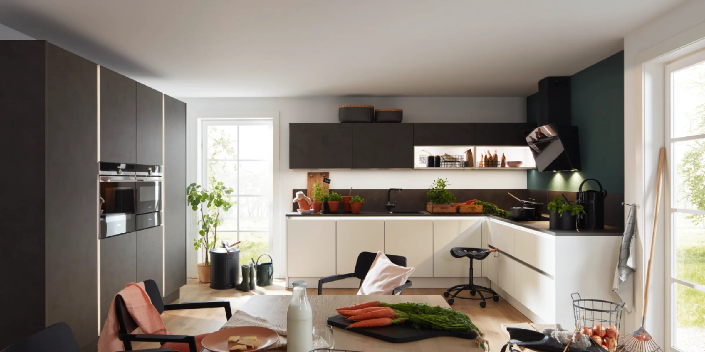 Moderne Nolte Küche mit der markanten Front PORTLAND in Zement Anthrazit und der Antifingerprint-Front PLUS in Weiß softmatt. Die zweifarbige Küchenzeile kombiniert dunkle Oberschränke mit hellen Unterschränken und bietet eine grifflose, pflegeleichte Gestaltung. Großzügige Fenster, Holzdetails und natürliche Deko-Elemente schaffen eine wohnliche, lichtdurchflutete Atmosphäre.