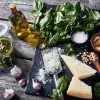 Auf einem Holztisch liegen Zutaten für Pesto, darunter Basilikum, Parmesan und Knoblauch.