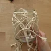 Eine Hand hält ein mit Makramee verziertes Glas auf einem Holztisch.