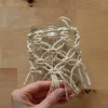 Eine Hand hält ein Glas mit Makramee-Verzierung auf einem Holztisch.