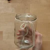 Eine Hand hält ein Glas mit einer Schnur darum auf einem Holztisch.