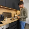 Ein Mann bereitet in einer modernen Küche mit Holz- und Schwarzakzenten Kaffee zu. Nolte Küchen.