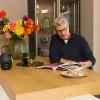 Ein Mann sitzt am Küchentresen mit Blumen und Makronen, während er ein Magazin liest. Nolte Küchen.