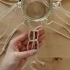 Eine Hand knotet Makramee-Fäden um ein Glas auf einem Holztisch.