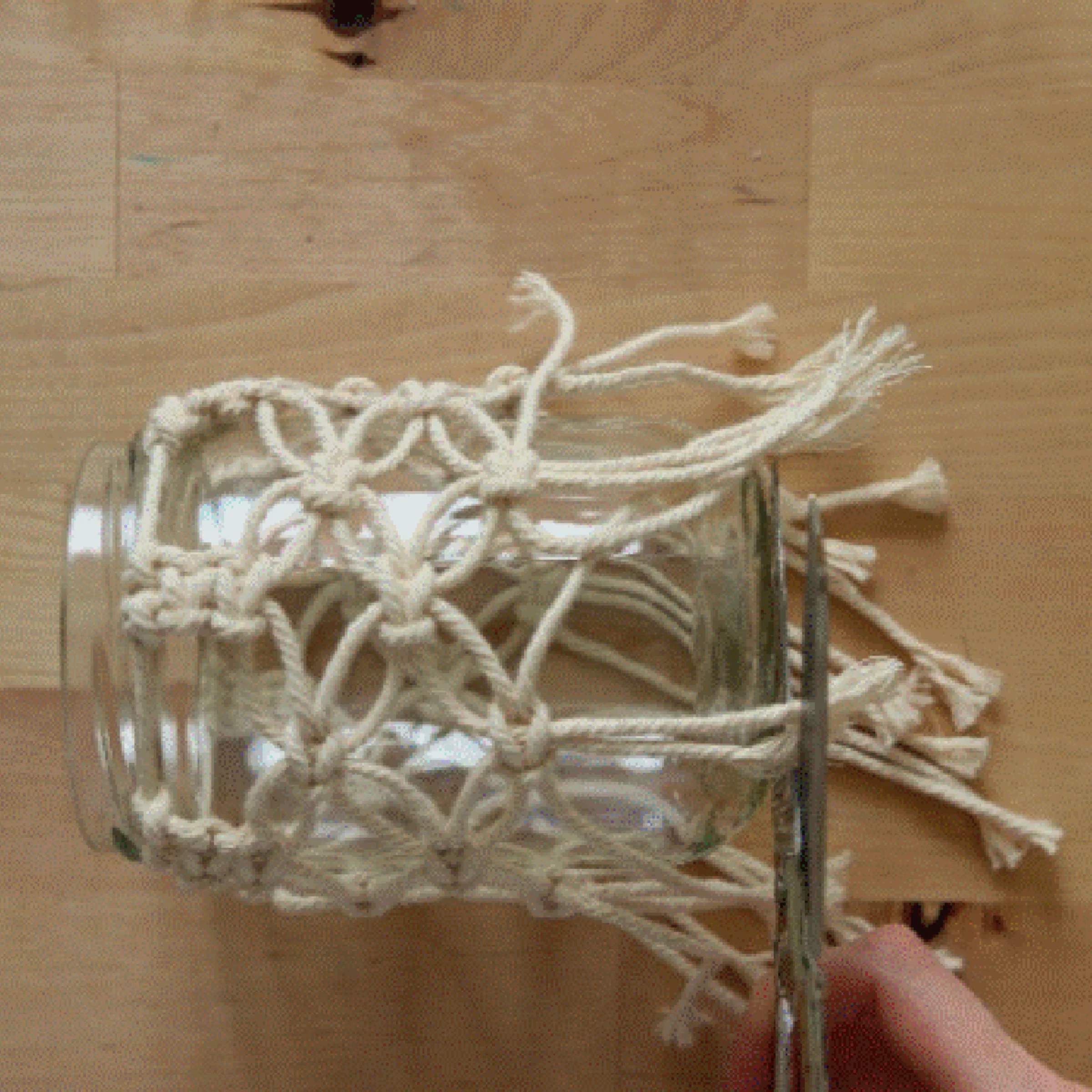 Ein Glas wird mit einem Makramee-Netz umwickelt, während jemand die Fäden abschneidet.
