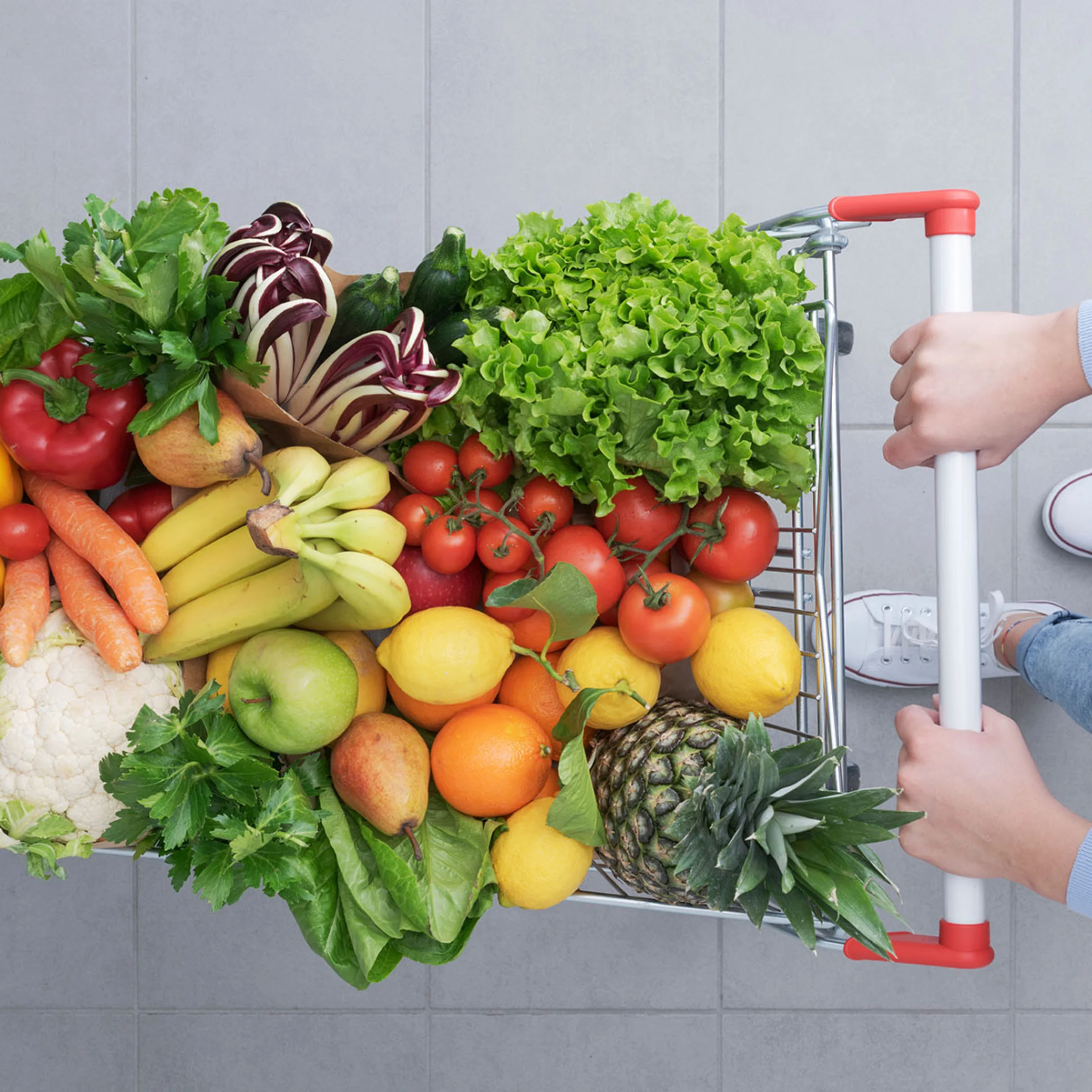 Eine Person schiebt einen Einkaufswagen voller frischem Obst und Gemüse auf einem grauen Fliesenboden.
