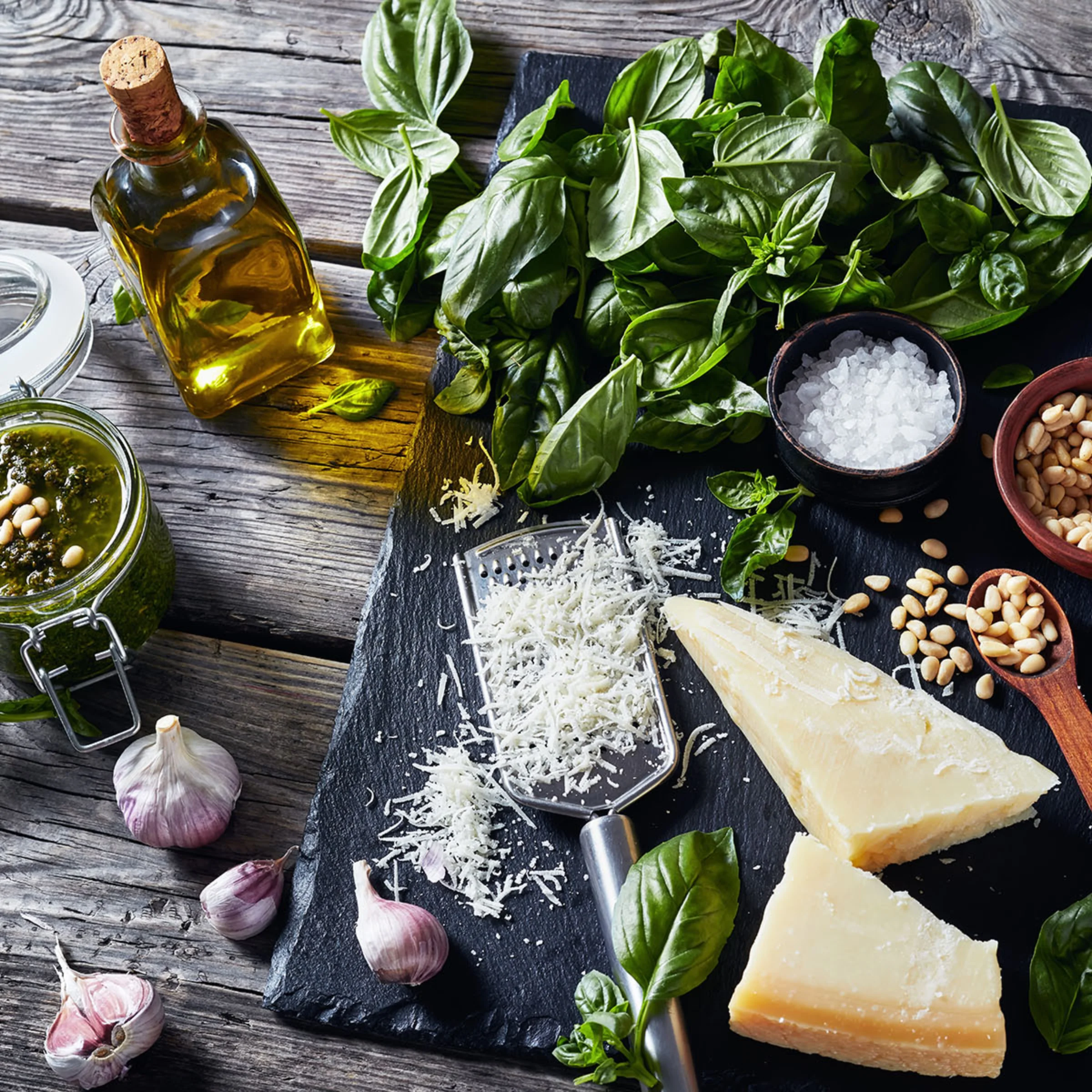 Auf einem Holztisch liegen Zutaten für Pesto, darunter Basilikum, Parmesan und Knoblauch.