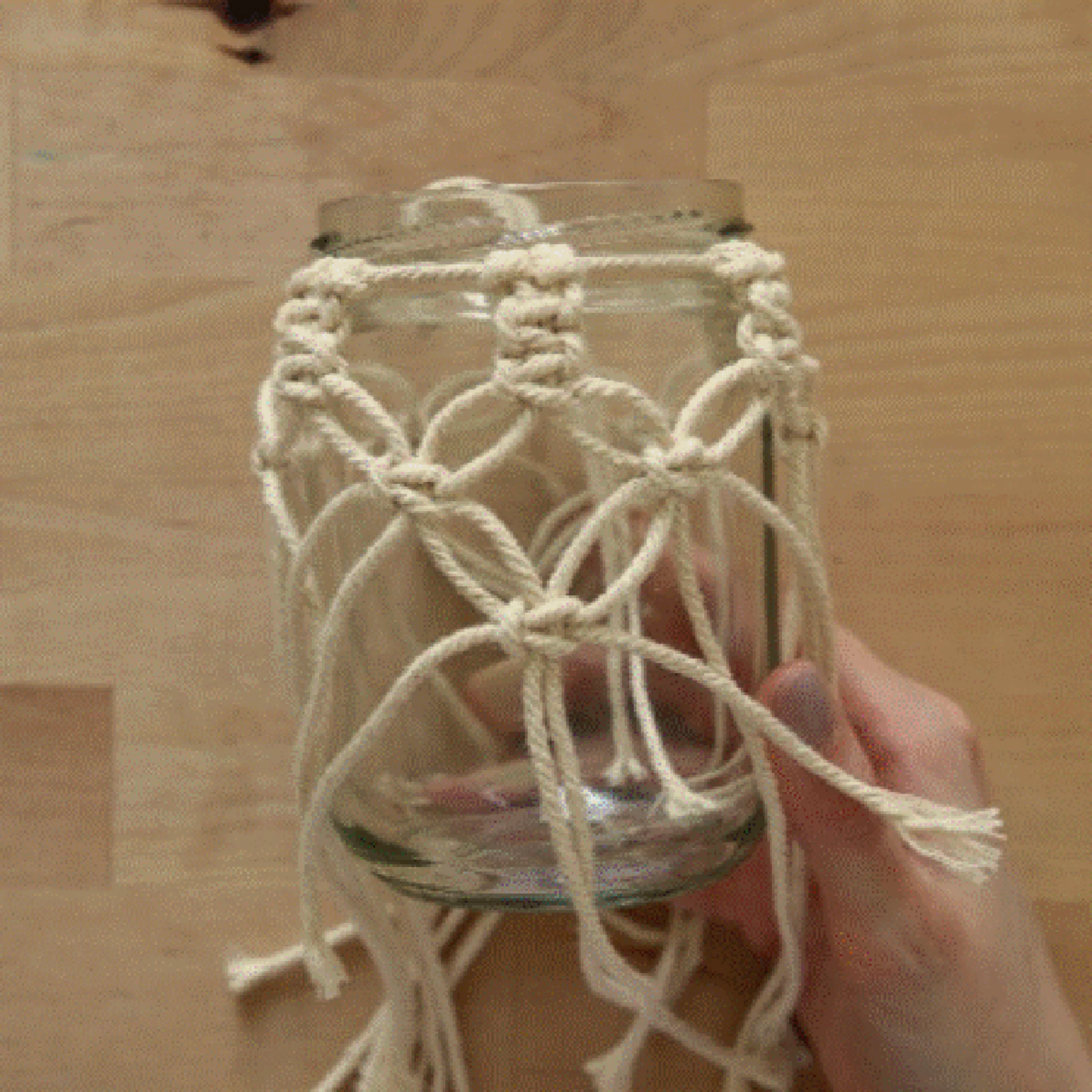 Eine Hand hält ein mit Makramee verziertes Glas auf einem Holztisch.