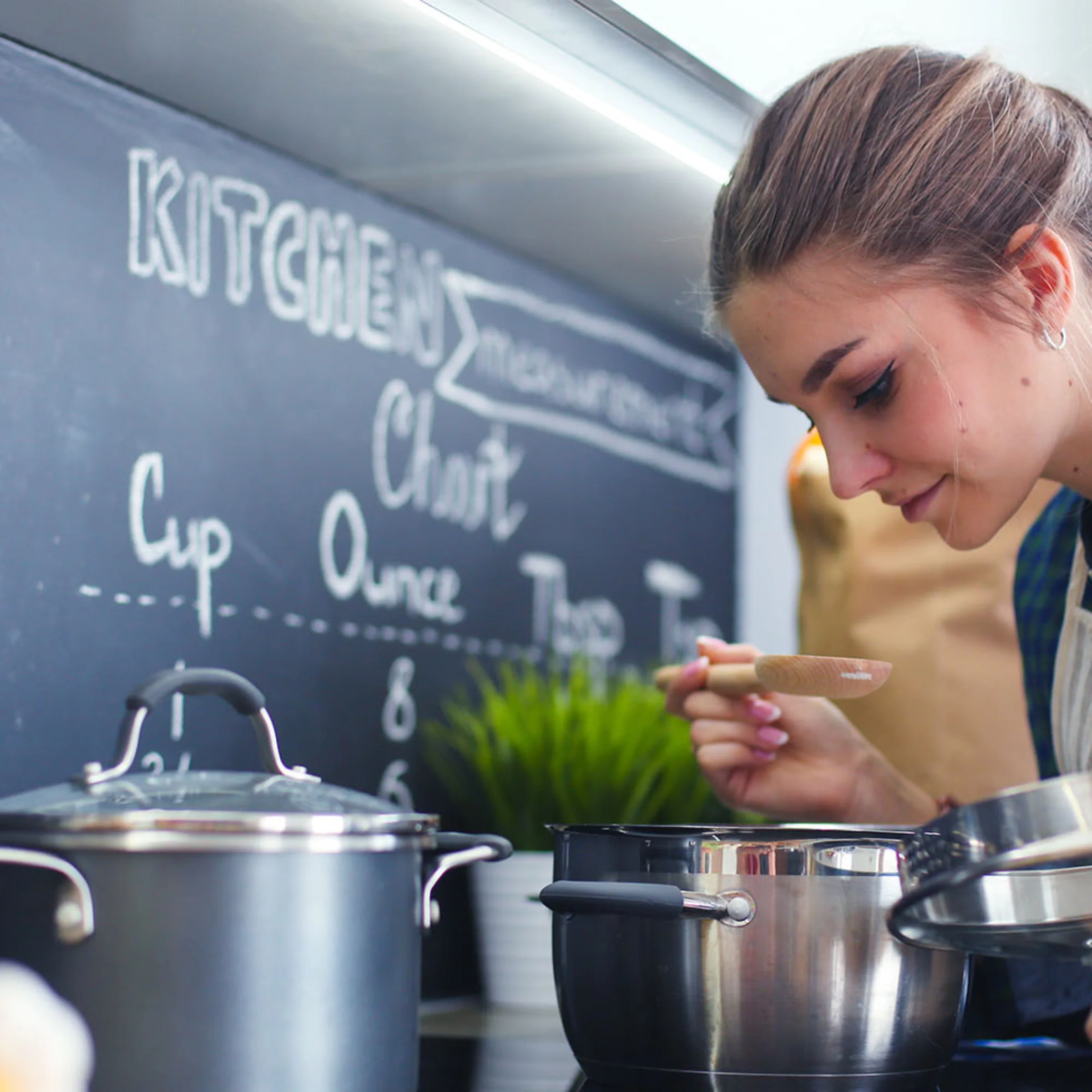 Eine Frau kocht in einer modernen Küche mit Töpfen und Kreidetafel im Hintergrund, Nolte Küchen.