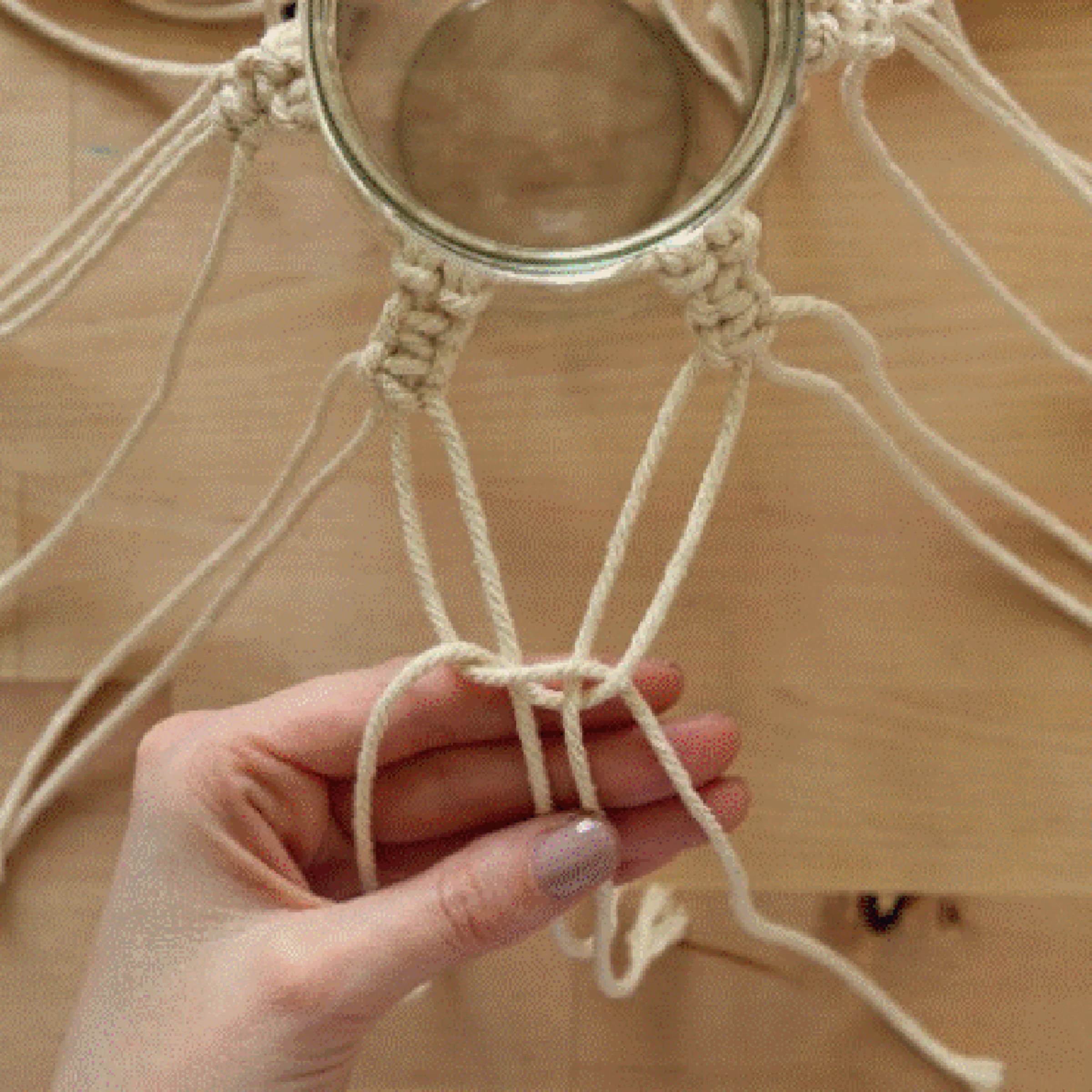 Eine Hand knotet Makramee-Seile um ein Glas auf einem Holztisch.