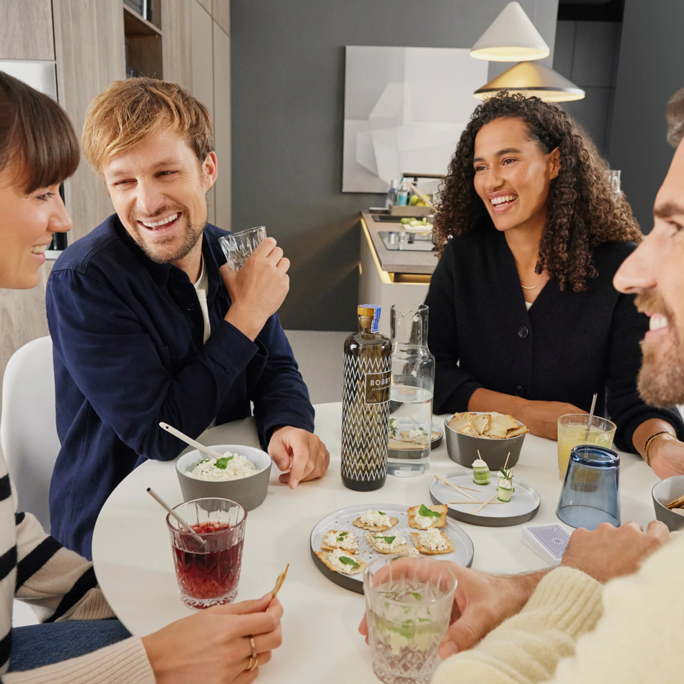 Vier Personen sitzen fröhlich an einem Tisch mit Getränken und Snacks in einem modernen Wohnbereich. Nolte Living.