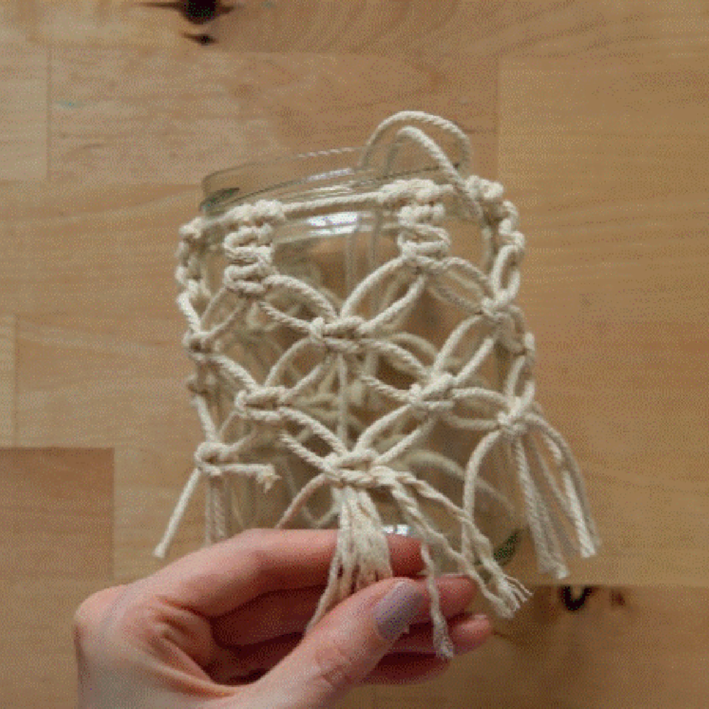 Eine Hand hält ein Glas mit Makramee-Verzierung auf einem Holztisch.