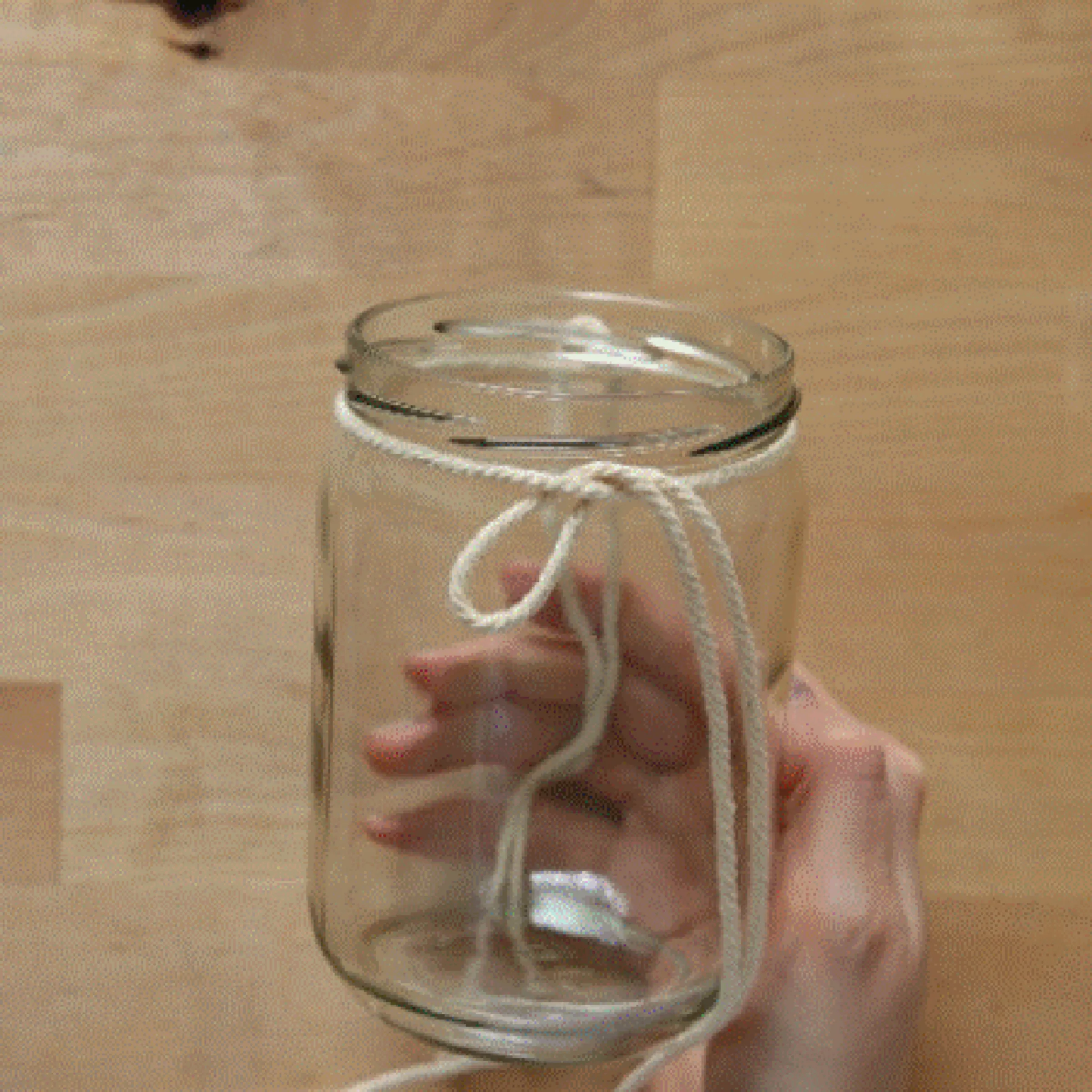 Eine Hand hält ein Glas mit einer Schnur darum auf einem Holztisch.