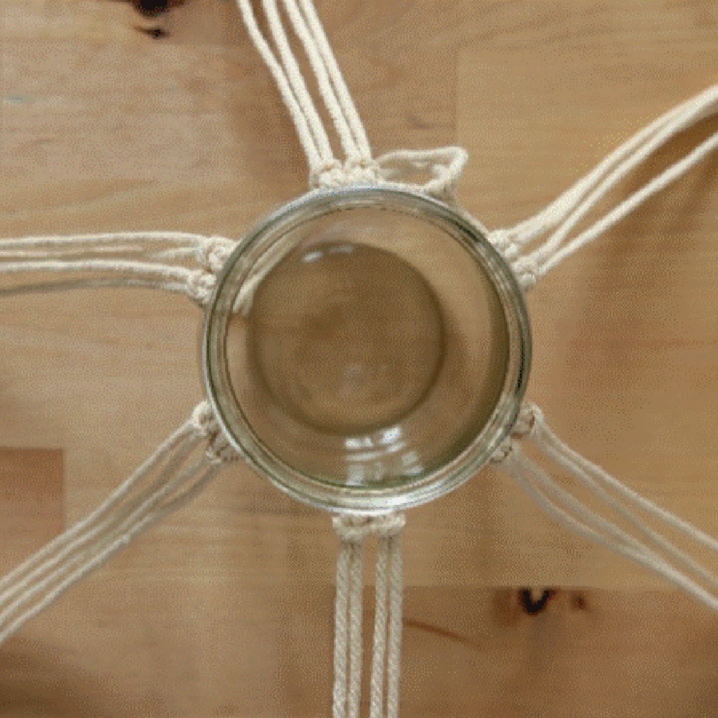 Ein Glas wird von Makramee-Schnüren gehalten, die auf einem Holztisch ausgebreitet sind.