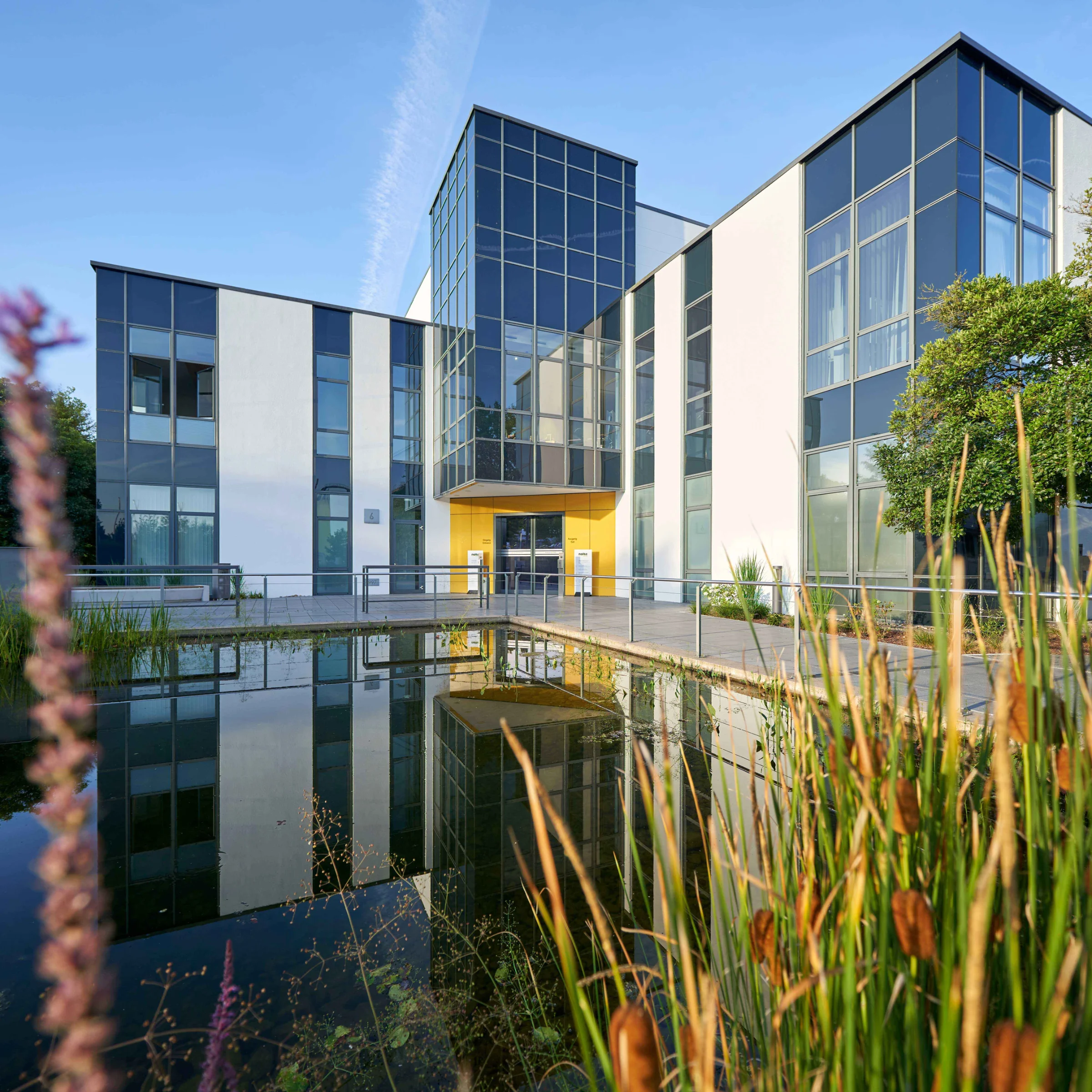 A modern glass-fronted building reflects in a pond, surrounded by plants. Nolte Kitchens.