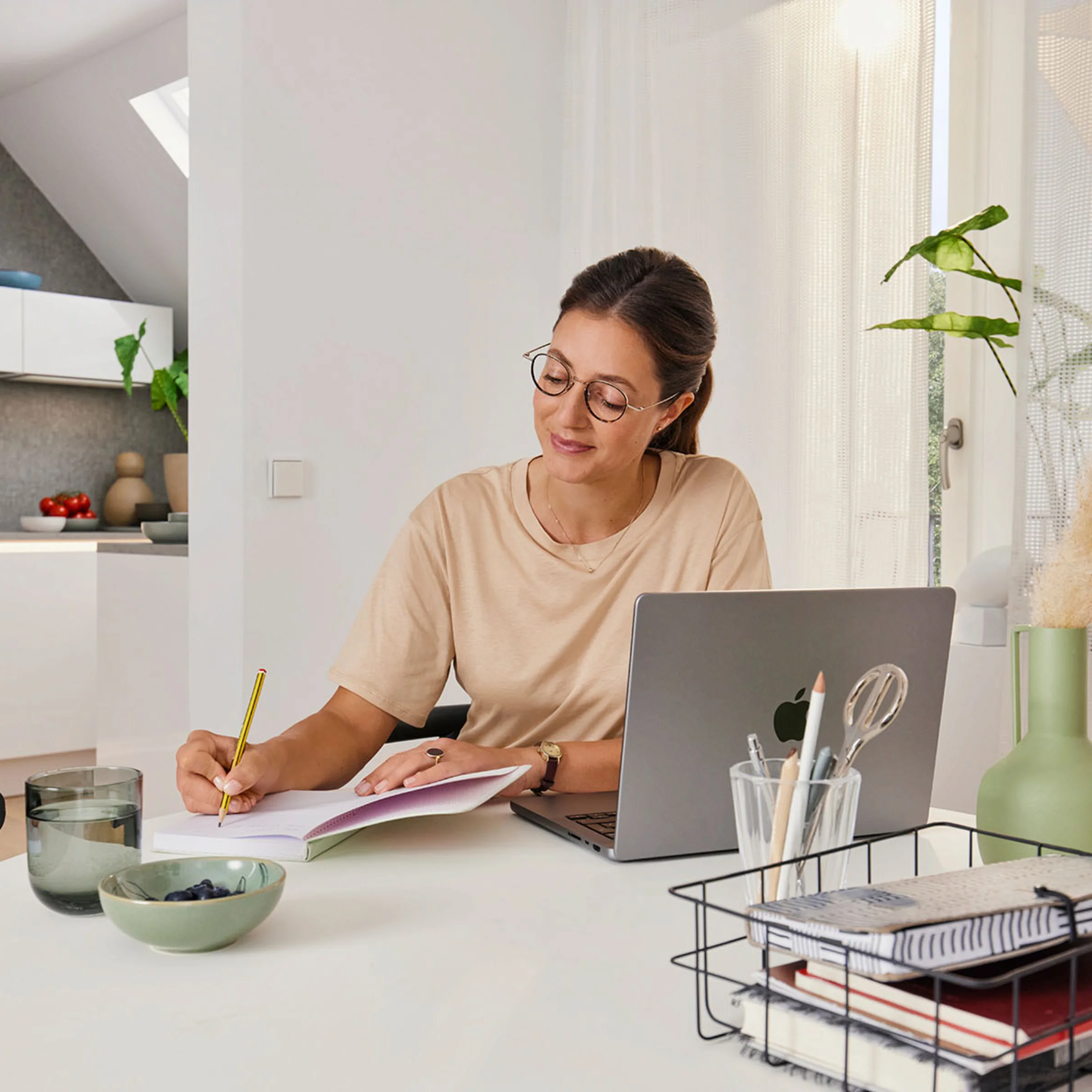 Eine Frau arbeitet an einem Tisch in einem modernen Raum mit offener Küche im Hintergrund von Nolte Küchen.