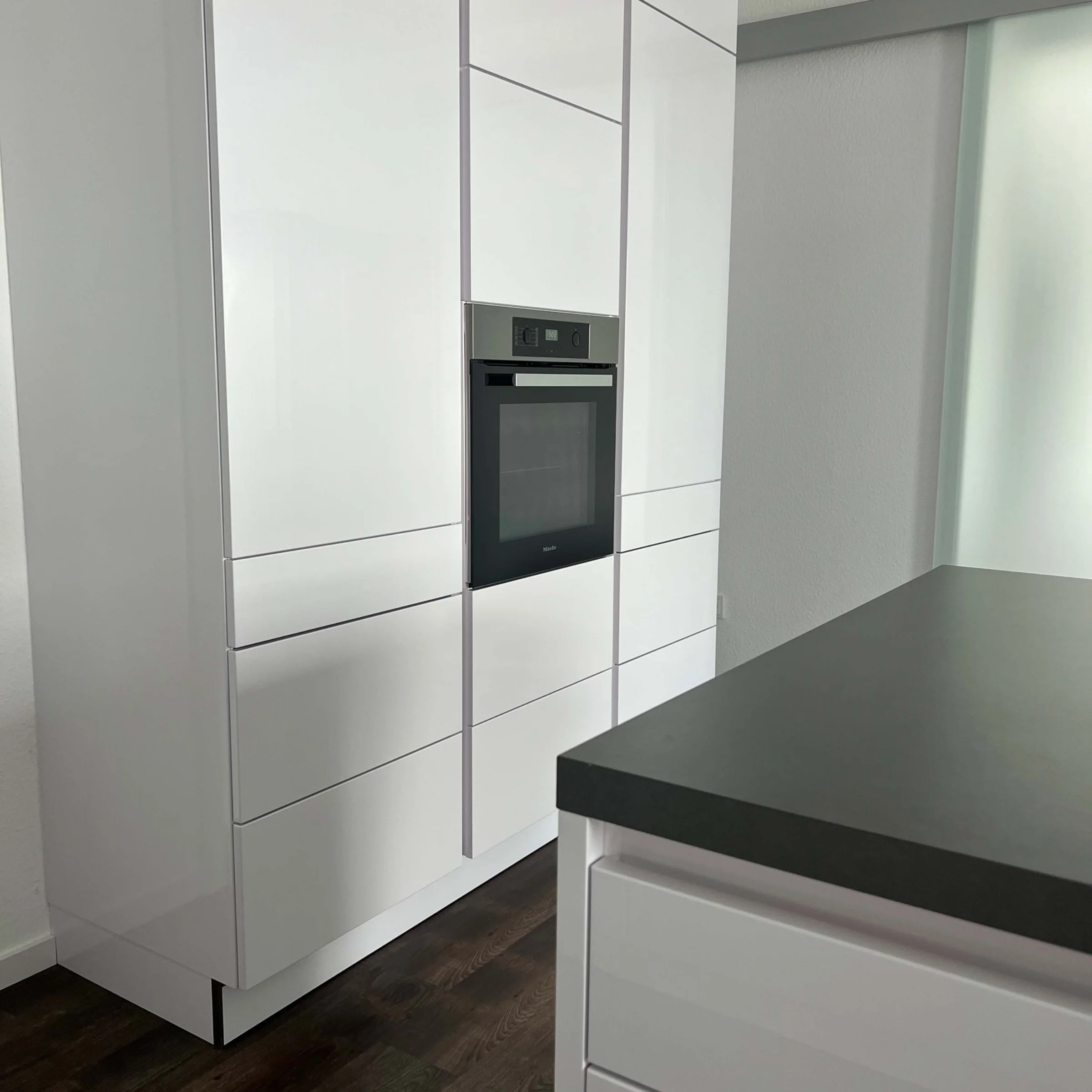 Eine moderne, weiße Küche mit eingebautem Ofen und dunklem Holzboden, gestaltet von Nolte Küchen.