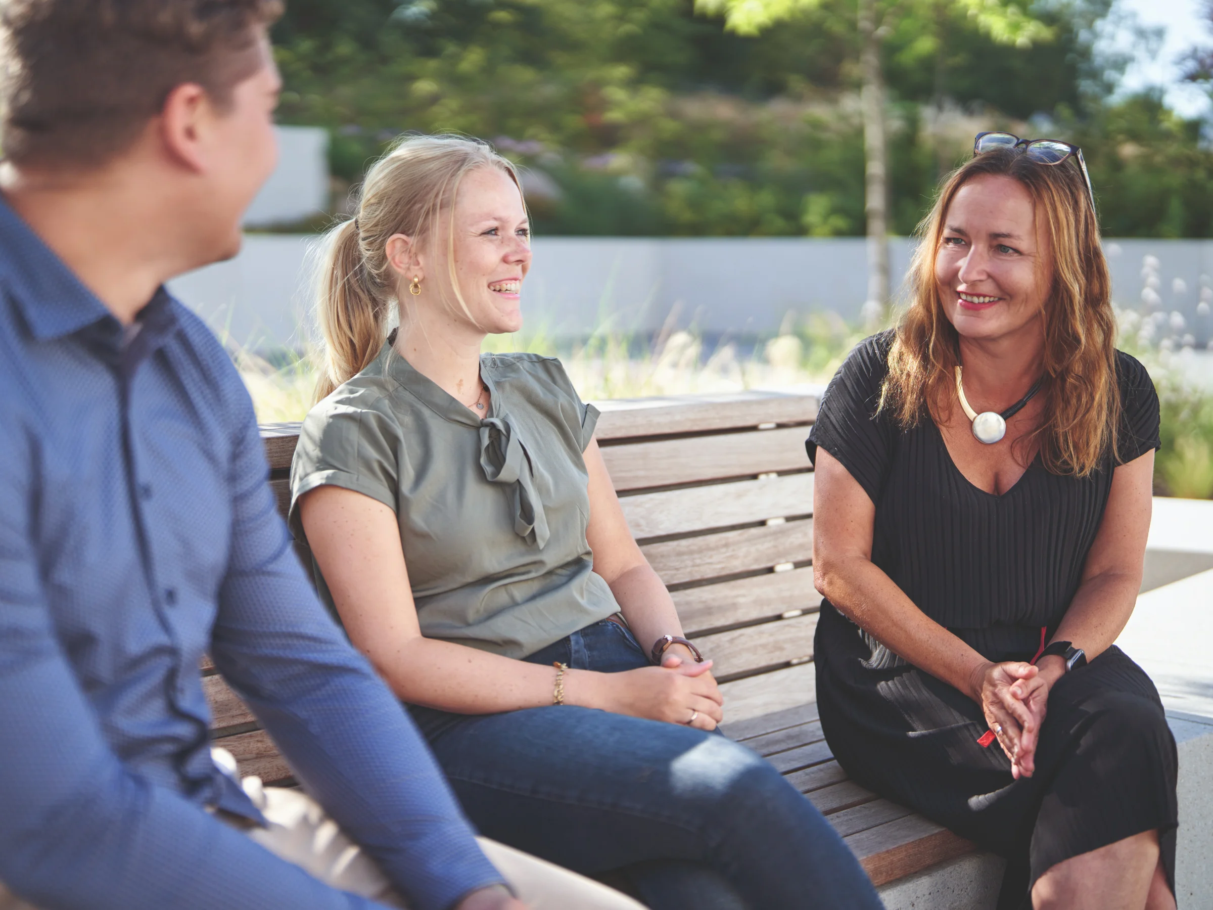 Tres personas sentadas en un banco al aire libre conversan bajo el sol.