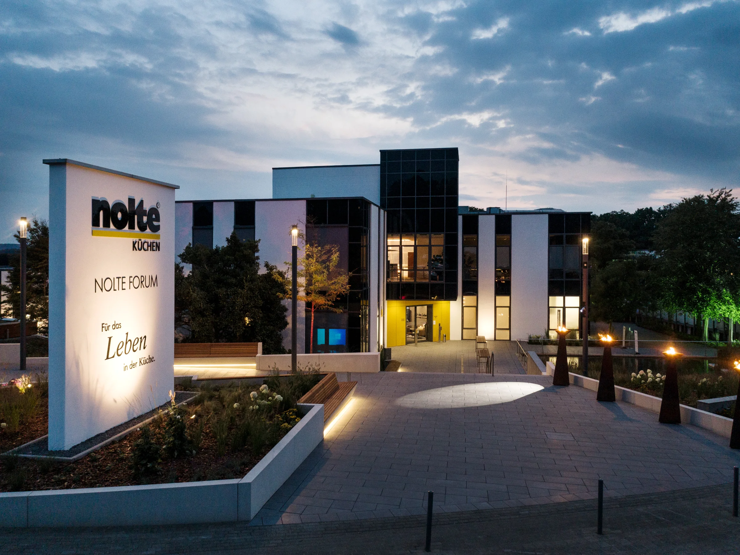 Ein modernes Gebäude bei Dämmerung mit beleuchtetem Nolte Küchen Schild im Vordergrund.