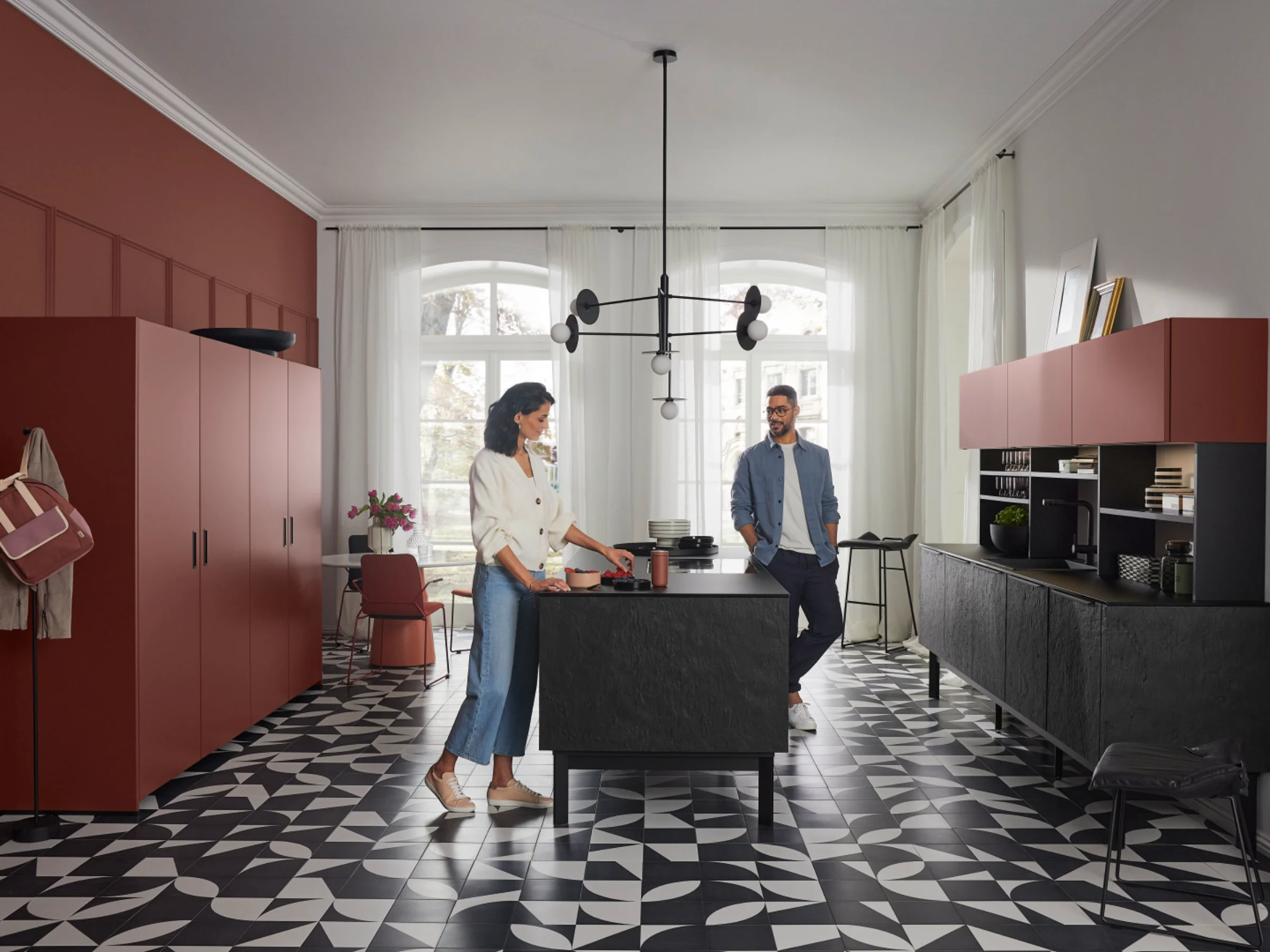 Eine moderne Küche von Nolte Küchen mit zwei Personen, geometrischem Fliesenboden und roten Schränken.