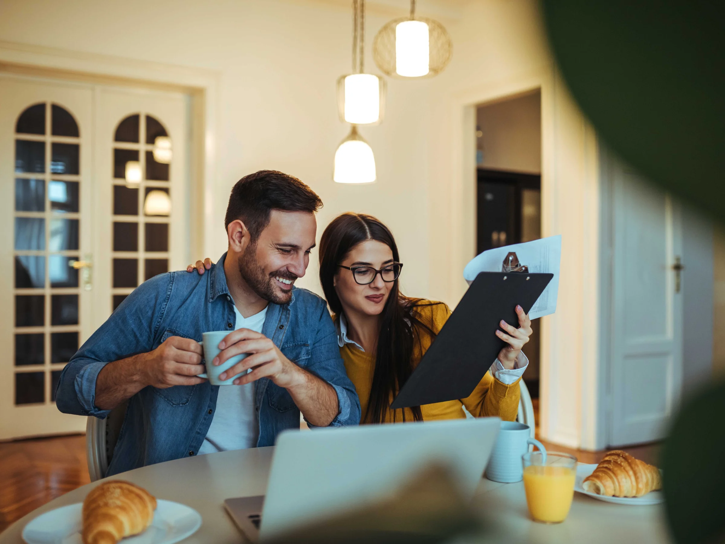 Ein Paar sitzt am Tisch beim Frühstück mit Laptop und Dokumenten.