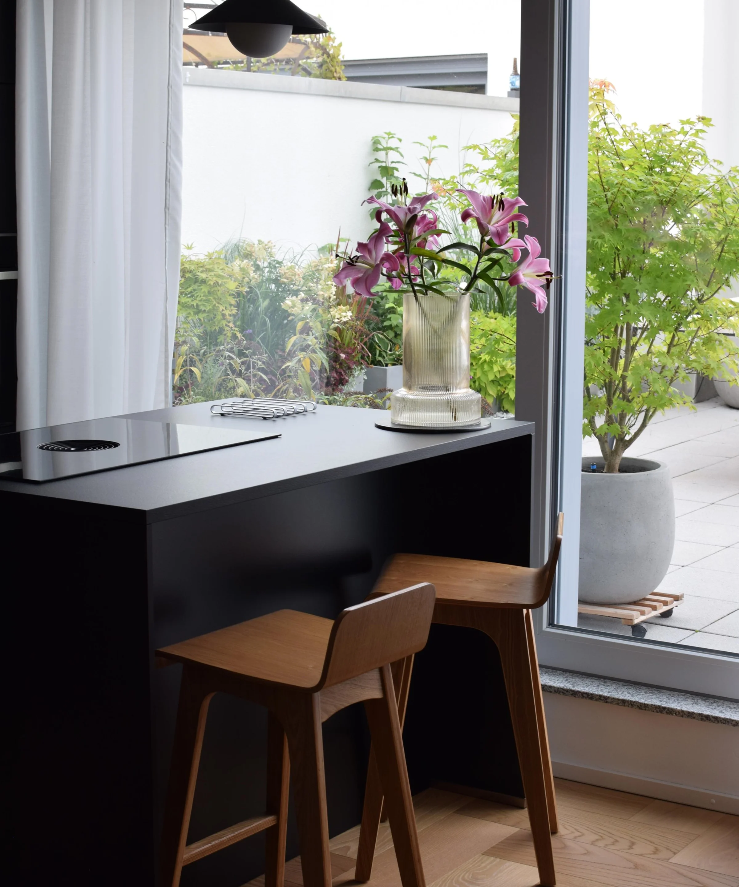 In a modern kitchen with large windows, two wooden chairs sit at a black counter. Nolte Kitchens.