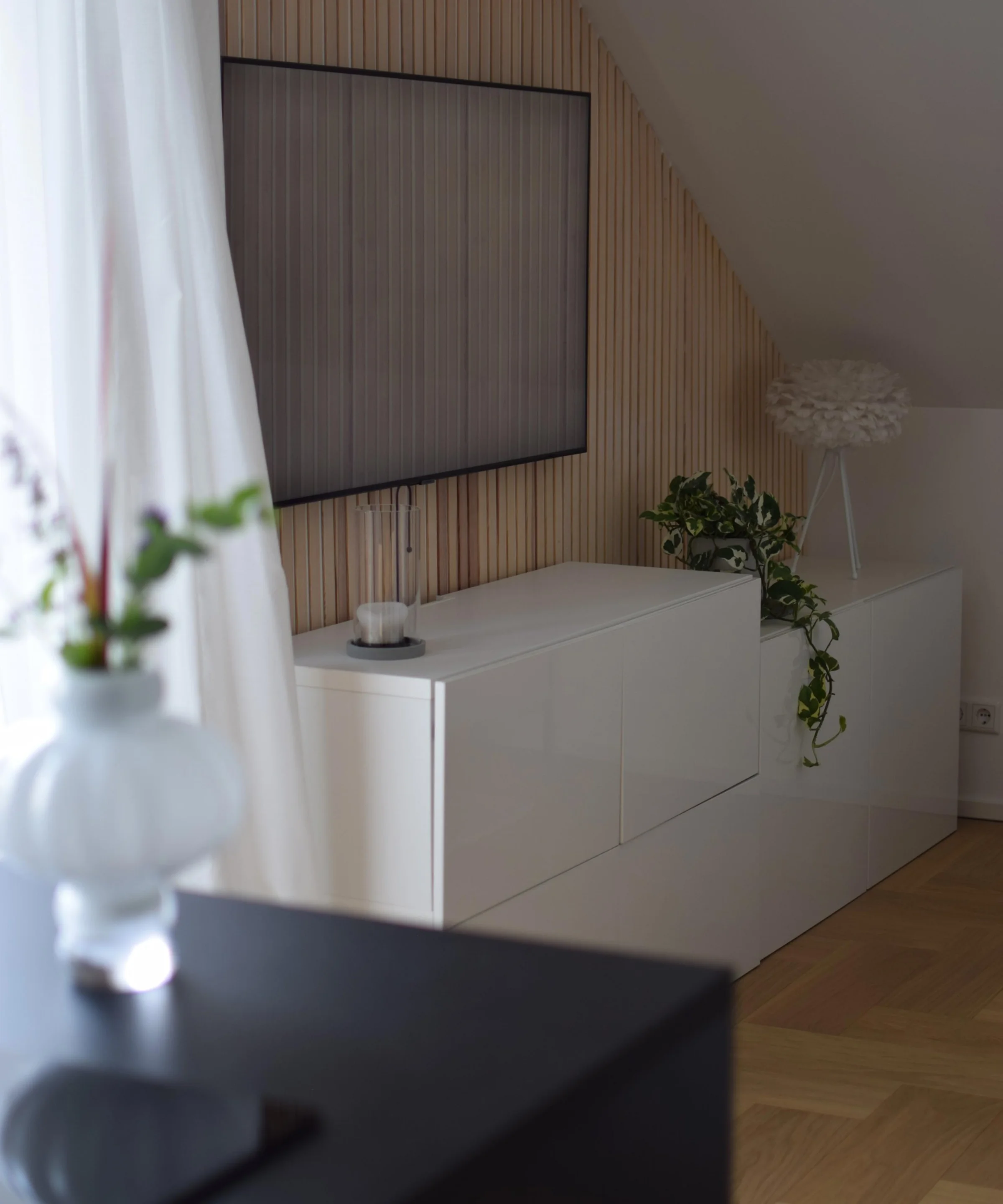 In einem modernen Wohnzimmer steht ein weißes Sideboard, dekoriert mit Pflanzen, unter einem Fernseher an der Wand. Nolte Living.